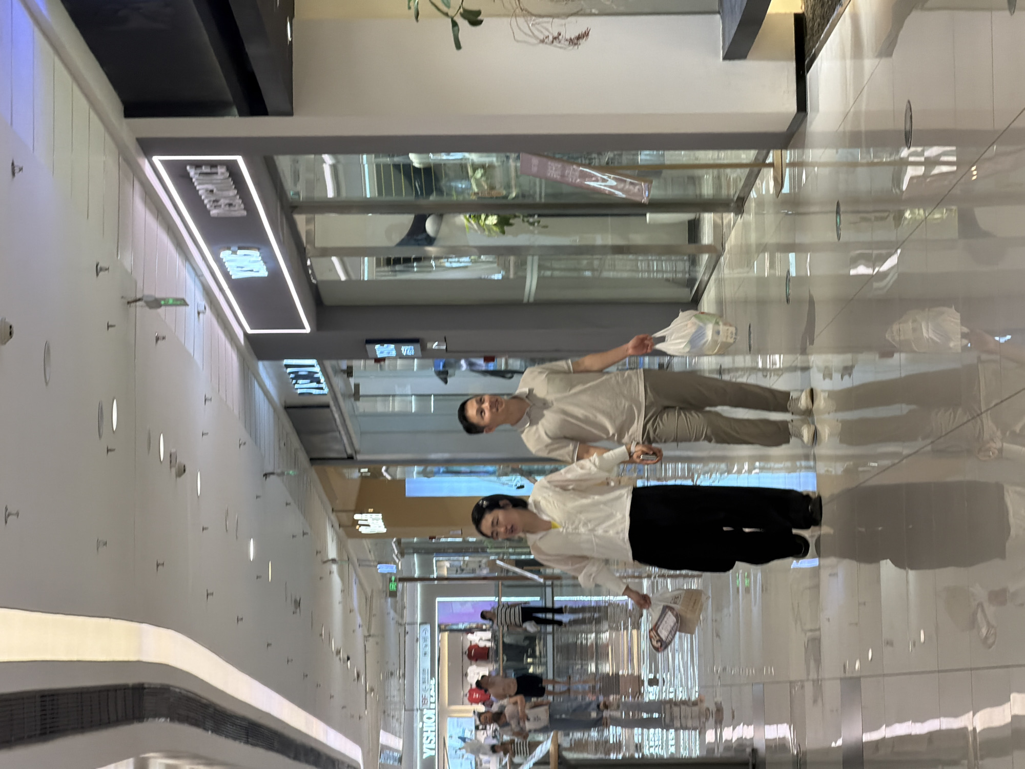 The image shows an indoor shopping mall in Jiangjin, China. Two people, a man and a woman, are walking towards the viewer. The woman is on the left, wearing a white long-sleeved shirt and black pants. She is carrying a shopping bag in her right hand. The man is on the right, wearing a light-colored polo shirt and light-colored pants. He is also carrying a shopping bag in his right hand. They are walking on a shiny tiled floor, which reflects the overhead lights and the surrounding environment. The mall has a modern design with a high ceiling and architectural elements. In the background, there are storefronts with illuminated signage, including one that reads "YSHION" and another with neon lights forming the words "YING DESIGNICE". Other shoppers can be seen in the distance. The overall atmosphere suggests a typical shopping scene.