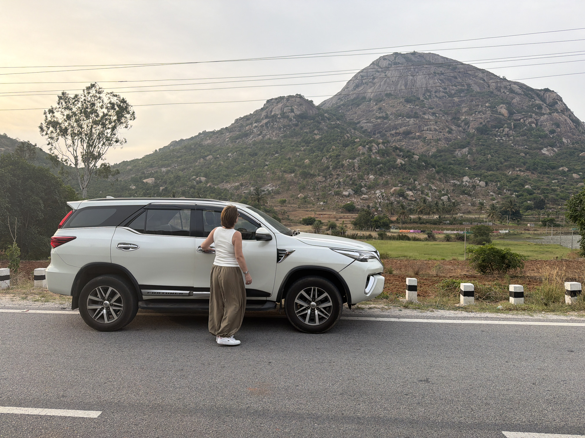 A woman stands by a white SUV parked on an asphalt road in Muddenahalli, India. The woman wears a white tank top, loose brown pants, and white sneakers. She rests her hand on the car door. The SUV has dark-tinted windows and alloy wheels. The road is marked by small white and black posts along the side. In the background, a large, rocky mountain dominates the horizon. Vegetation and a few scattered trees dot the landscape around the mountain. The sky is overcast.
