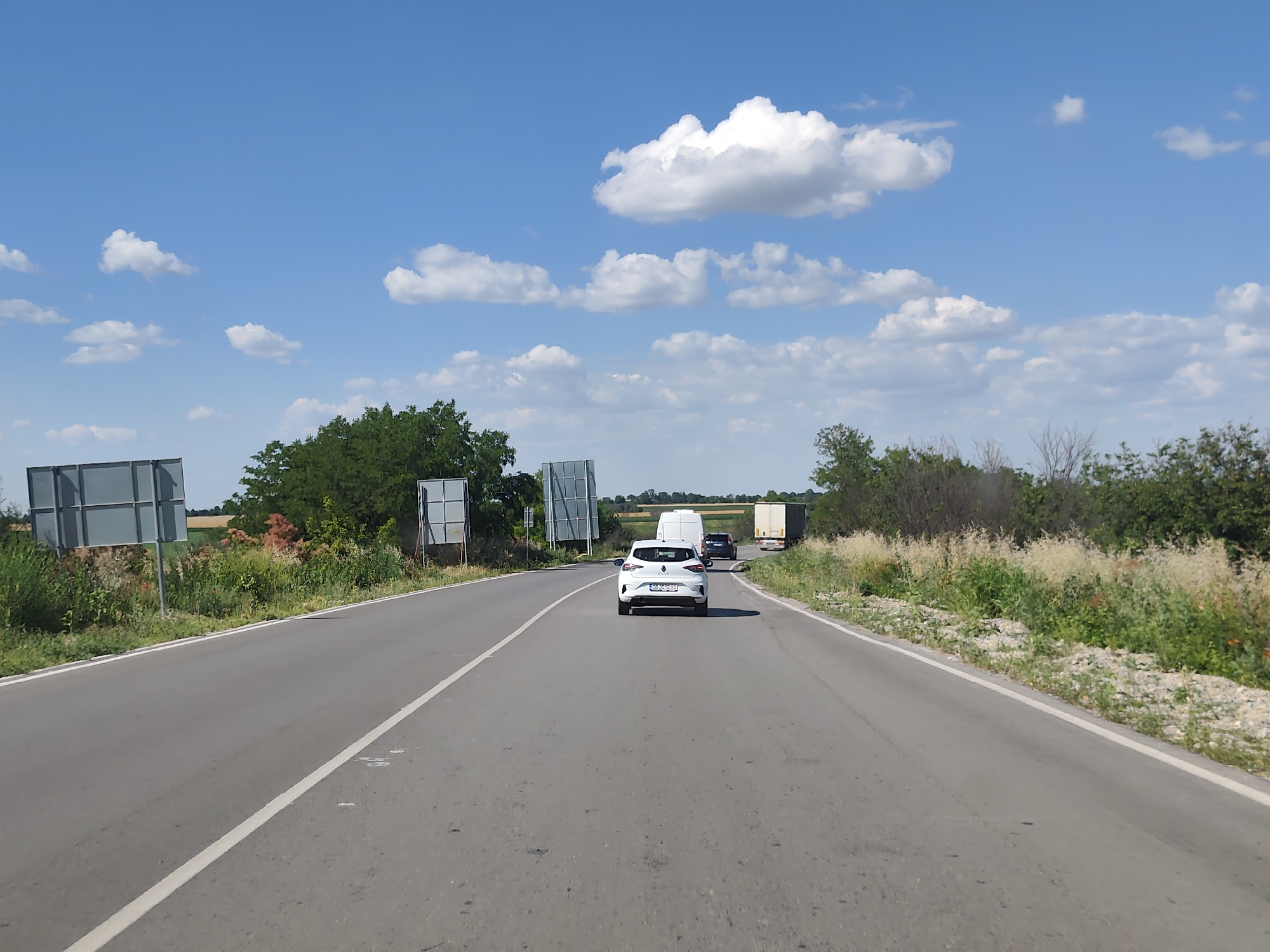 The image depicts a sunny day on a rural road in Yasen, Bulgaria. The road is paved and has white lane markings. On the left side of the road, there are two large, rectangular signs with grey surfaces. Beyond the signs, agricultural fields and trees are visible. On the right side of the road, there is dense vegetation, including trees and bushes, with some lighter colored plants in the foreground.
Several vehicles are on the road, traveling in the same direction. The closest vehicle to the viewer is a white car, which appears to be a hatchback. Further ahead, there is a dark-colored car, followed by a white van and a large truck with a white trailer. The activity taking place is travel on a highway or main road.
The sky is a vibrant blue with several fluffy white clouds scattered across it, indicating pleasant weather. The sunlight suggests it is daytime, likely late morning or early afternoon. The overall scene is one of a typical drive through a countryside environment. The license plate of the white car is partially visible and appears to have Cyrillic characters. The signs on the left side of the road are likely informational or directional, though their content is not legible from this image.