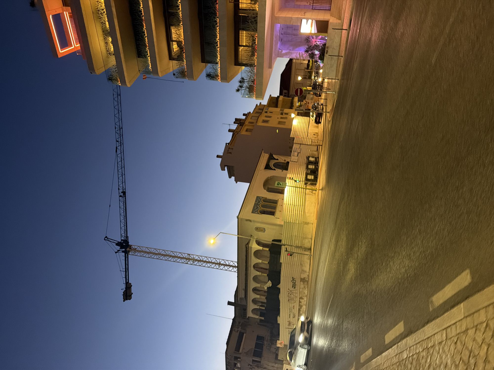 The image captures a street scene in Cascais, Portugal at dusk or early night. A tall construction crane dominates the upper left portion of the frame, silhouetted against a clear, deep blue sky. To the right, a modern, multi-story building is illuminated by warm, orange lights, highlighting its balconies adorned with decorative elements.
In the mid-ground, a street runs from left to right. On the left side of the street, a silver sedan is driving away from the camera, its headlights on. Further down the street, a series of buildings with arched windows and traditional architecture are visible. A streetlamp with a yellow glow illuminates the scene. Construction fencing lines the side of the road in front of these buildings.
The road surface appears wet and reflective, suggesting recent rain or street cleaning. White dashed lines mark the lanes on the road. The sidewalk on the left is paved with cobblestones. Several cars are parked or driving on the street in the distance, and some buildings in the background are also lit. The overall impression is an urban environment at twilight, with a mix of old and new architecture and ongoing construction.