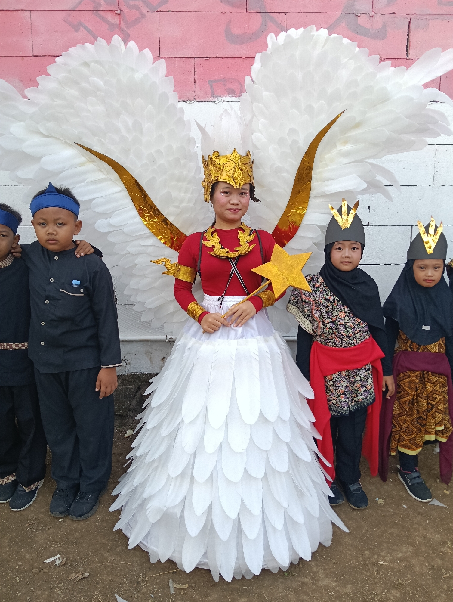In Tasikmalaya, Indonesia, a group of children stand for a photograph in front of a white brick wall adorned with pink graffiti. The central figure, a young person, wears a striking white feathered costume with large golden wings and a golden crown. They hold a gold star-shaped wand. Flanking this central figure are four children dressed in traditional Indonesian attire. To the left, two boys wear dark blue uniforms and headbands. To the right, two children wear black headscarves, one with a vibrant patterned garment and red sash, and the other with a patterned skirt. The ground is a dirt surface with some scattered debris.