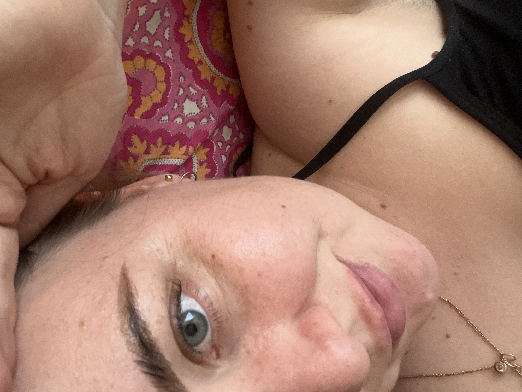 The image presents a close-up, high-angle shot of a woman lying down in an indoor setting in Chaikovsky, Russia. Her face is prominently featured in the foreground, turned to her right, with her gaze directed towards the camera. She has light skin with numerous freckles visible across her nose and cheeks. Her left eye, which is light blue or green, is open and clear. Her dark eyebrows are well-defined. Her lips are slightly parted, revealing a glimpse of her tongue, suggesting a relaxed posture. She wears a small gold stud earring in her earlobe and a delicate gold hoop in her upper ear cartilage.
In the upper right section of the frame, part of her body, likely her shoulder and upper chest, is visible. She is wearing a black top with a thin strap. A delicate gold-colored chain necklace with a small, circular charm, possibly containing a clear stone, rests on her skin. The background features a vibrant textile pattern, predominantly pink with intricate designs in red, orange, and yellow, indicative of a pillow or bedding. The lighting is soft and ambient, suggesting an indoor environment. The woman's expression appears neutral or slightly content. No visible text is present in the image.