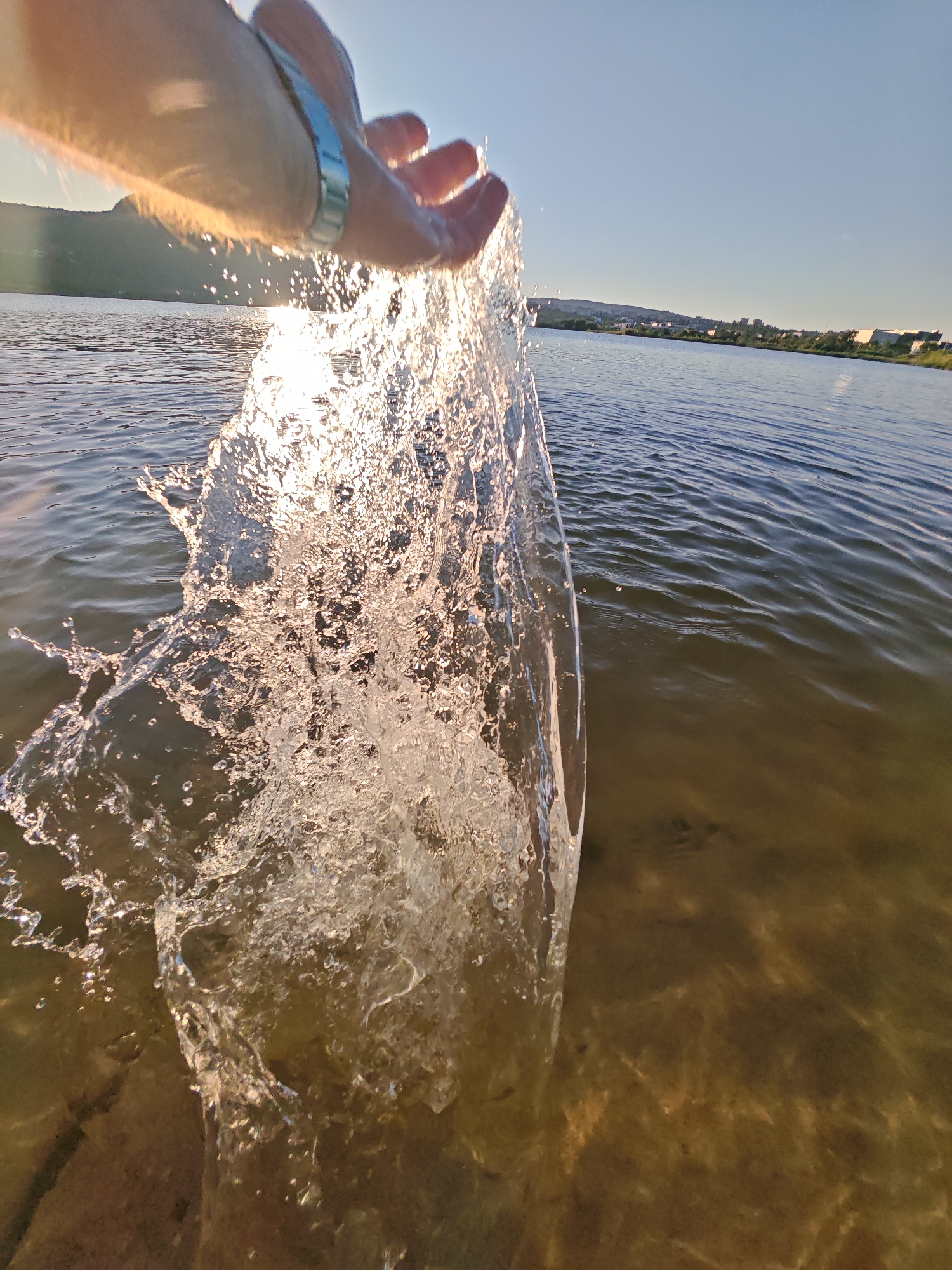The image shows a close-up, low-angle view of a person's hand splashing water in a lake or river. The sun is setting or rising, casting a warm glow on the water and the spray. The background shows a distant shoreline with buildings and trees, under a clear blue sky. The water is relatively calm with ripples, and the bottom of the lake is visible in the shallow areas, showing sand and some rocks. The person is wearing a silver bracelet on their left wrist. The overall atmosphere is serene and natural. The image was taken in the city of Shumen, Bulgaria.