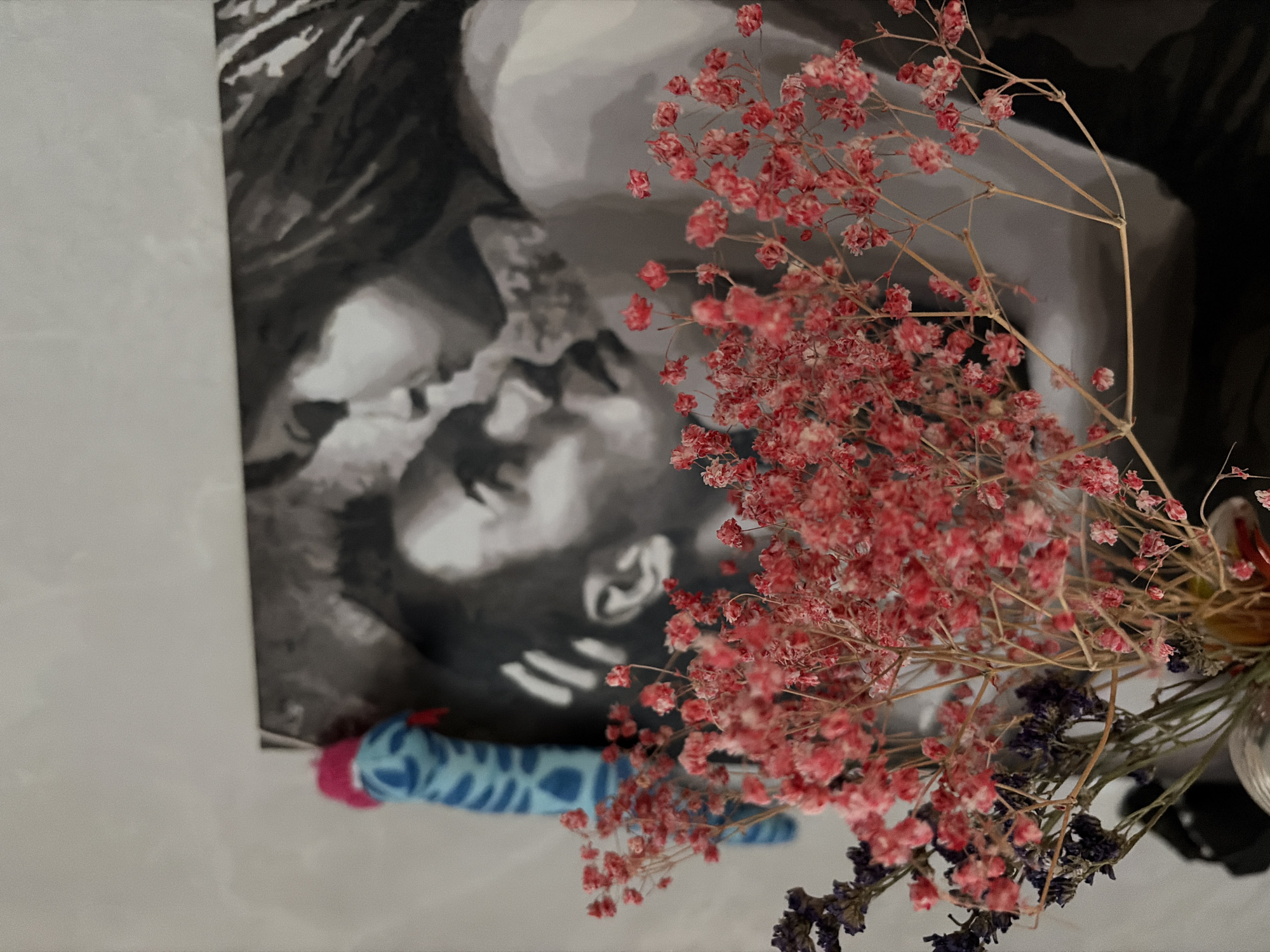 The image presents an indoor still life arrangement, dominated by a bouquet of dried flowers positioned in front of a black and white artwork. The main artwork, likely a painting or print, depicts a stylized, grayscale representation of a person's head and upper torso. The figure's head is tilted slightly, conveying a pensive or contemplative mood, with a soft focus or painterly effect on their features.
In the foreground, a prominent bouquet of dried flowers occupies the right side of the frame. These flowers are primarily a delicate, vibrant pink, resembling dried baby's breath (Gypsophila), and are clustered densely. Interspersed among them are some darker, dried purple flowers, possibly lavender, adding a touch of color variation. The stems of the flowers are visible, leading down to a partially obscured clear glass or vase holding them on the far right.
At the bottom left, a small, cylindrical object with a blue and white patterned fabric and a visible pink end is partially in view. The background consists of a light-colored, possibly white or off-white, surface extending to the left and behind the artwork.
No specific activity or event is taking place, and there is no visible text within the image. The setting appears to be indoors, making it impossible to determine the time of day or weather. While no visual cues within the image indicate a specific geographical location, the provided context states it is Perm, Russia. The overall impression is one of gentle beauty and quiet introspection.