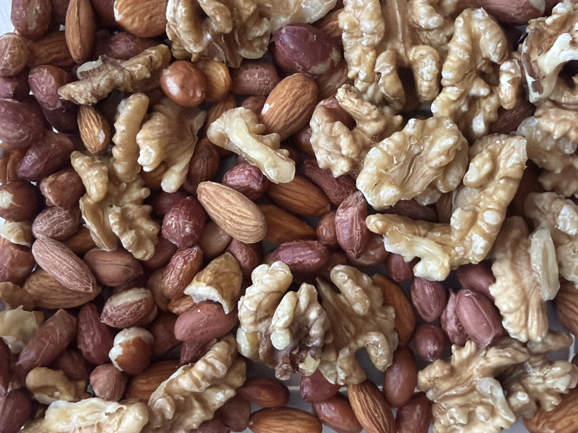 The image shows a close-up of a mix of nuts. The nuts present are walnuts, almonds, and peanuts. The walnuts are shelled and appear as irregular, pale brown pieces. The almonds are whole, brown, and elongated. The peanuts are also whole, reddish-brown, and rounded. The nuts are scattered randomly and densely, filling the entire frame. No people, subjects, actions, interactions, or location are discernible.