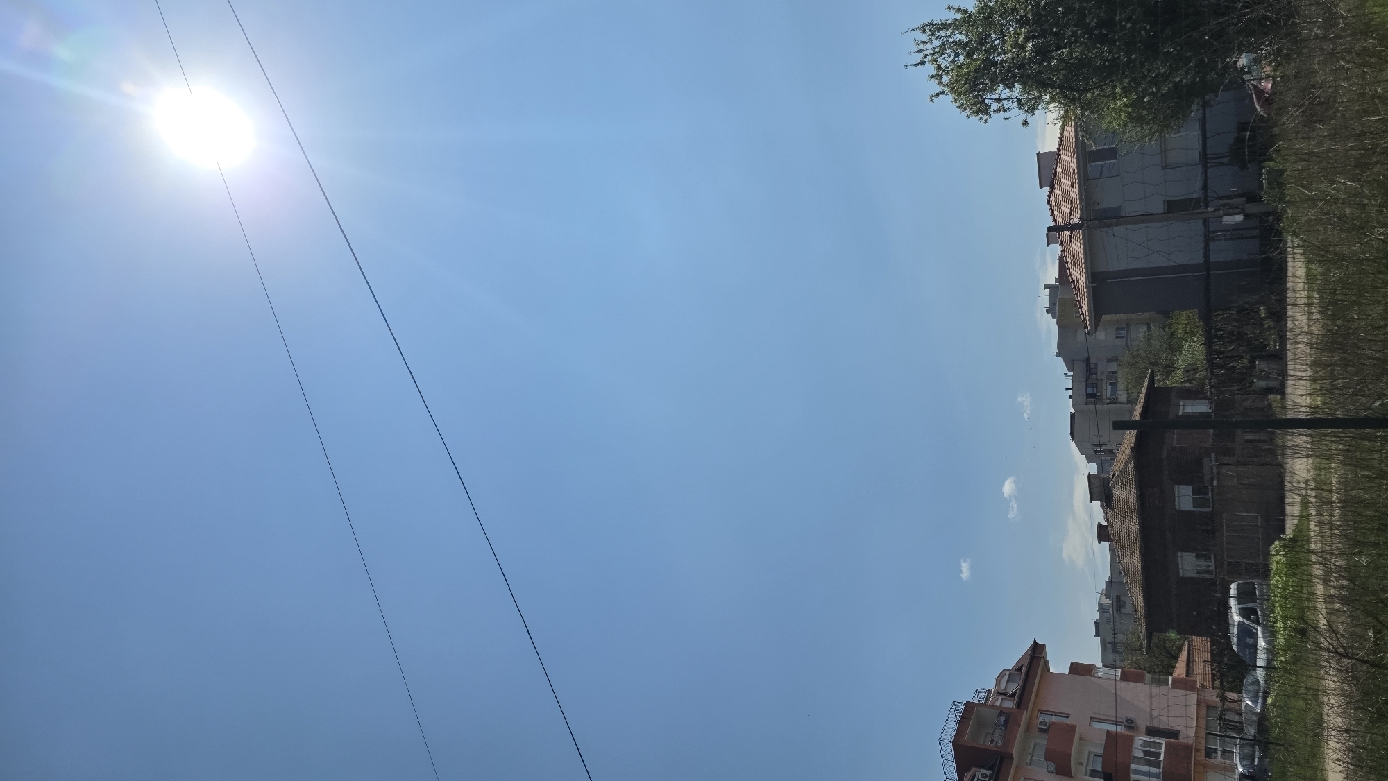 The image captures a cityscape under a bright, cloudless, blue sky. The sun is visible in the upper left corner. Power lines run diagonally across the left side of the frame. On the right side, there are several buildings, some appearing to be residential, with a mix of architectural styles. A building under construction with scaffolding and unpainted walls is visible. Trees are visible at the top and right of the image. The photo appears to be taken at ground level, looking upwards at an angle. The specific location in Blagoevgrad, Bulgaria, cannot be determined solely from the provided image.