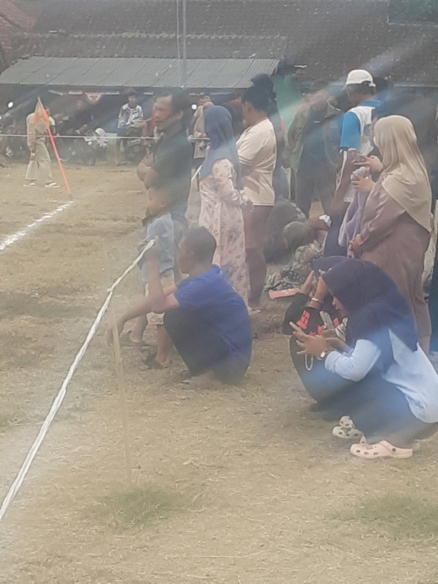 In the city of Cibalong, Indonesia, an outdoor event is taking place. The image shows a gathering of people, some of whom are watching an activity on a grassy field marked with a white line. In the background, a row of structures with corrugated roofs suggests a temporary event setup.
A person dressed in a light-colored outfit and a conical hat stands on the left, holding a red flag attached to a pole. Nearby, a child in a blue shirt is being held by an adult who is partially obscured. Further to the right, a group of individuals are seated or crouched on the ground, observing the event. One person, wearing a dark blue shirt, is kneeling and holding a wooden stick. Another person in the foreground is wearing a light blue long-sleeved shirt and dark pants, with a dark blue headscarf. This individual is looking down and appears to be interacting with a phone. A pair of pink clogs are visible on their feet. Other individuals in the middle ground are dressed in casual clothing, including a woman in a light-colored hijab and a woman in a patterned dress. The overall atmosphere seems to be one of community engagement at an outdoor event.