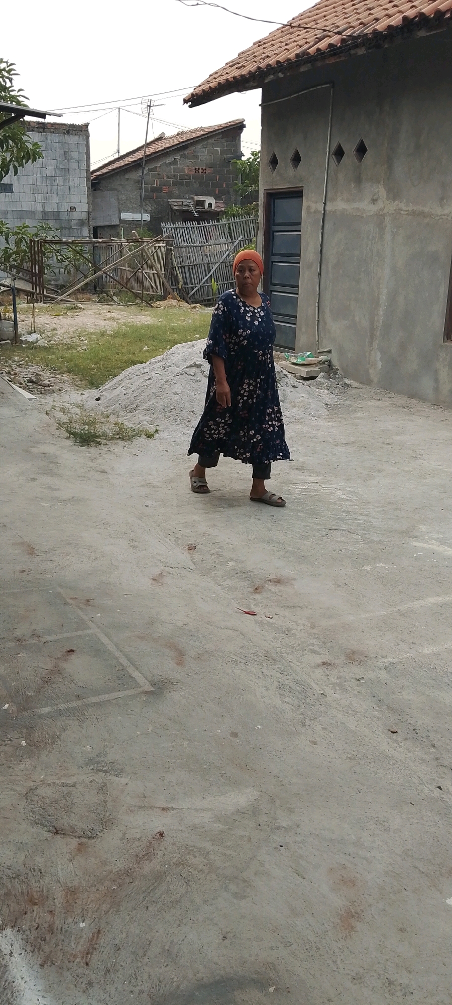 A person is walking on a dirt path in front of a building. The person is wearing a dark blue floral dress and an orange headscarf. There is a pile of sand to the left of the person and some buildings in the background. The scene appears to be in an outdoor, semi-urban environment in Sriamur, Indonesia.