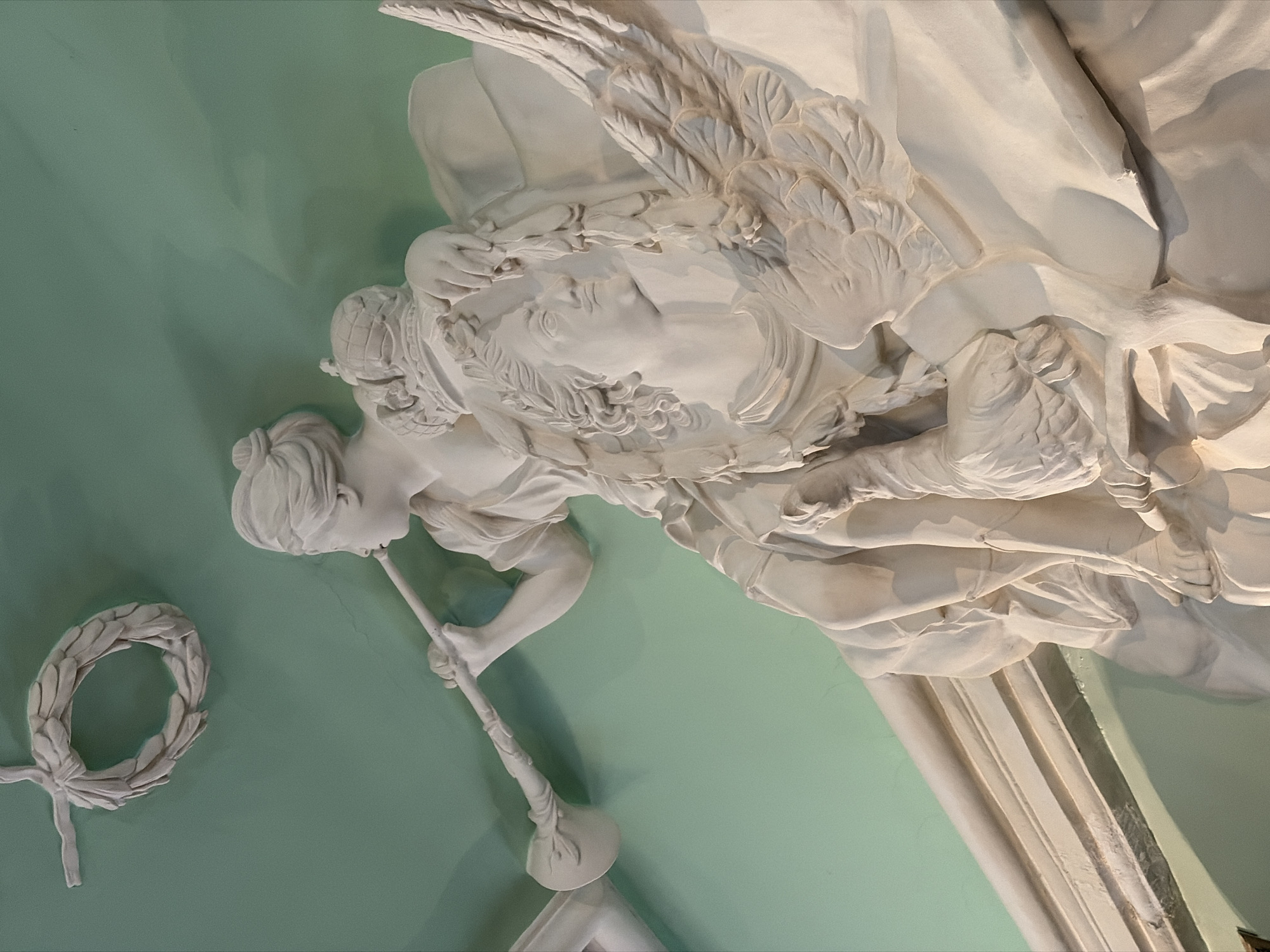 The image depicts a plaster relief sculpture against a light green background. The sculpture features a winged figure, possibly an angel or allegorical figure, blowing a long, slender trumpet. The figure has detailed features, including a laurel wreath on its head and flowing robes. To the left of the figure is a separate plaster wreath.
This relief is located in Saint Petersburg, Russia.