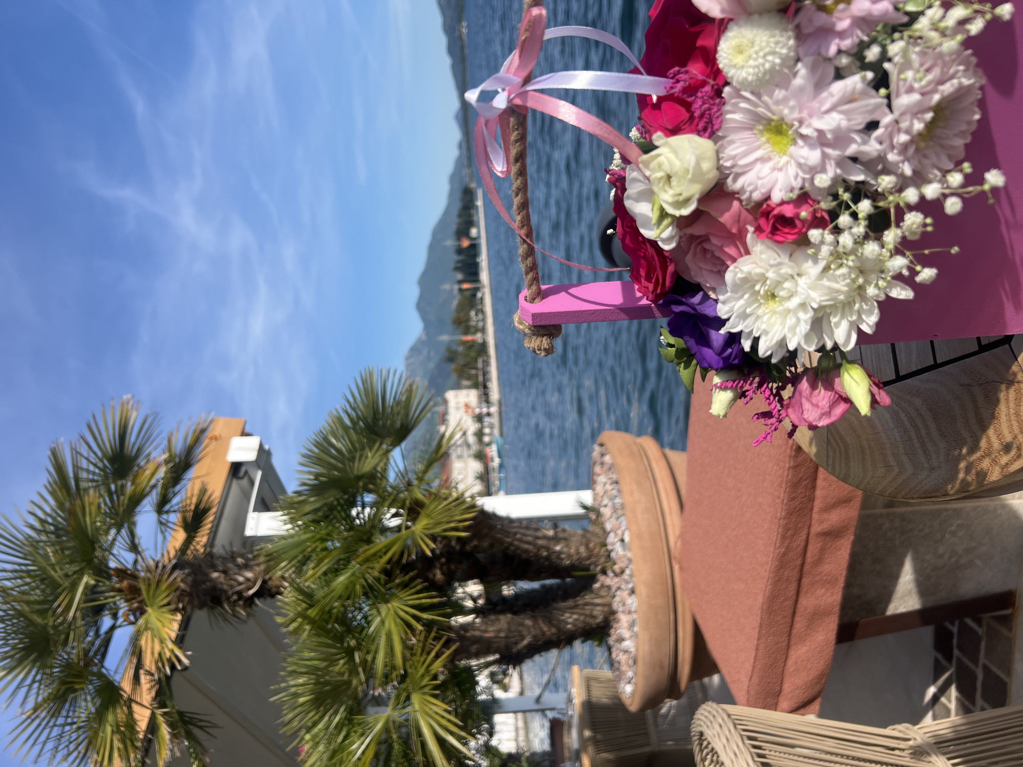 The photograph appears to be taken from a low angle, likely at an outdoor dining area in Tivat, Montenegro, possibly near the Porto Montenegro marina. In the foreground, there is a vibrant floral arrangement in a pink container hanging from a small, decorative pink swing suspended by rope with white and pink ribbons. The flowers consist of pink, white, and purple varieties, including roses and chrysanthemums. The background features a clear, blue sky and a wide expanse of water, suggesting the Adriatic Sea. Mountains are visible in the distance across the water, providing depth to the scene. Palm trees are present on the left side of the frame. The scene is tranquil and potentially related to an event or celebration, judging by the floral arrangement.