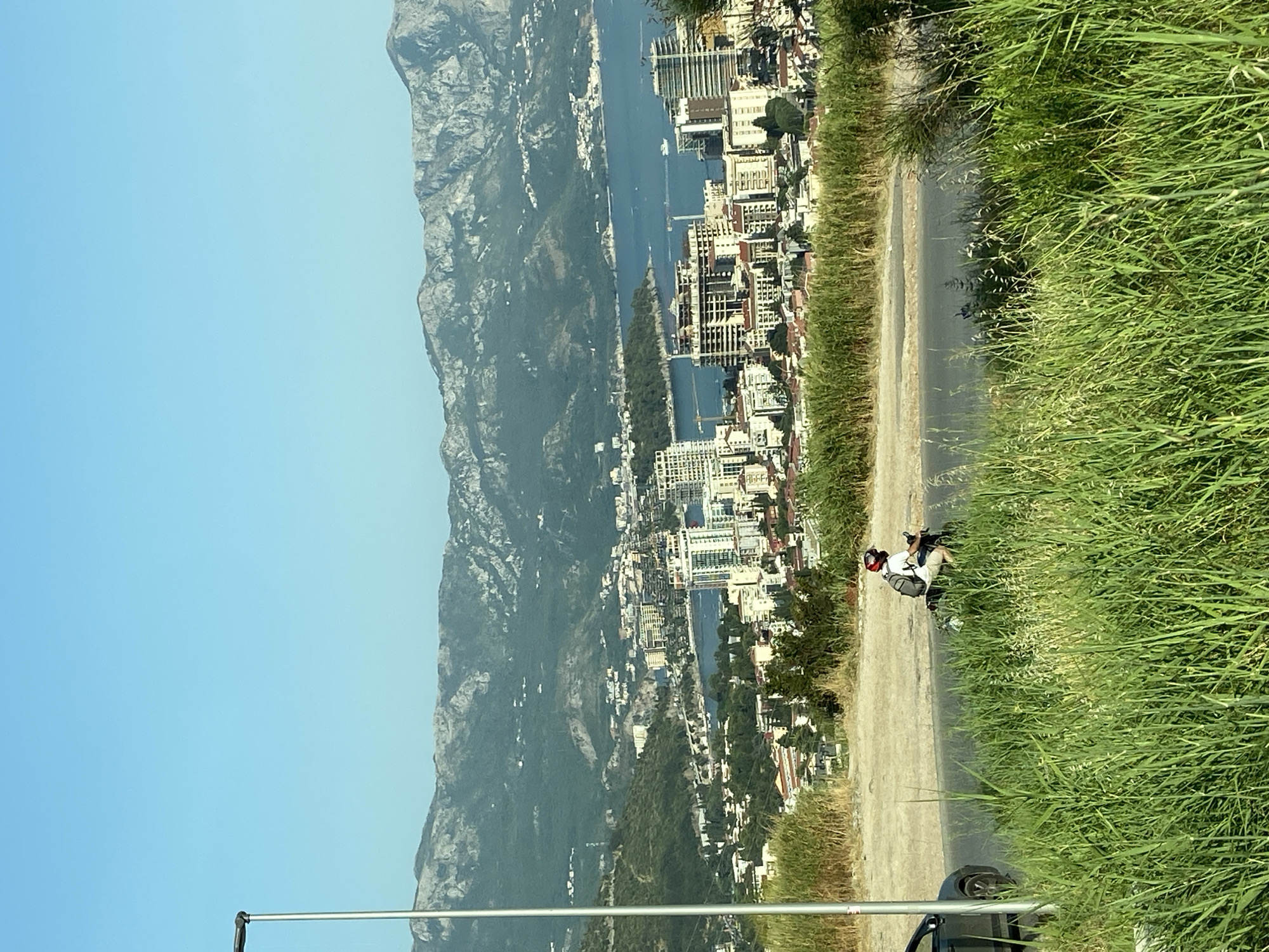 The image shows a panoramic view of a coastal city, likely Podostrog, Montenegro, with mountains in the background. In the foreground, lush green grass and tall reeds are visible. A paved road runs horizontally across the middle ground, with a person on a scooter riding away from the camera. To the left of the road, a dark-colored car is partially visible. Beyond the road, the city unfolds with buildings of various sizes and architectural styles, including modern high-rises and older structures with terracotta roofs. A bay or harbor is present, dotted with boats. The mountains in the distance are rugged and covered in green vegetation, with some patches of rock. The sky is a clear, bright blue. A street lamp is positioned on the left side of the image.