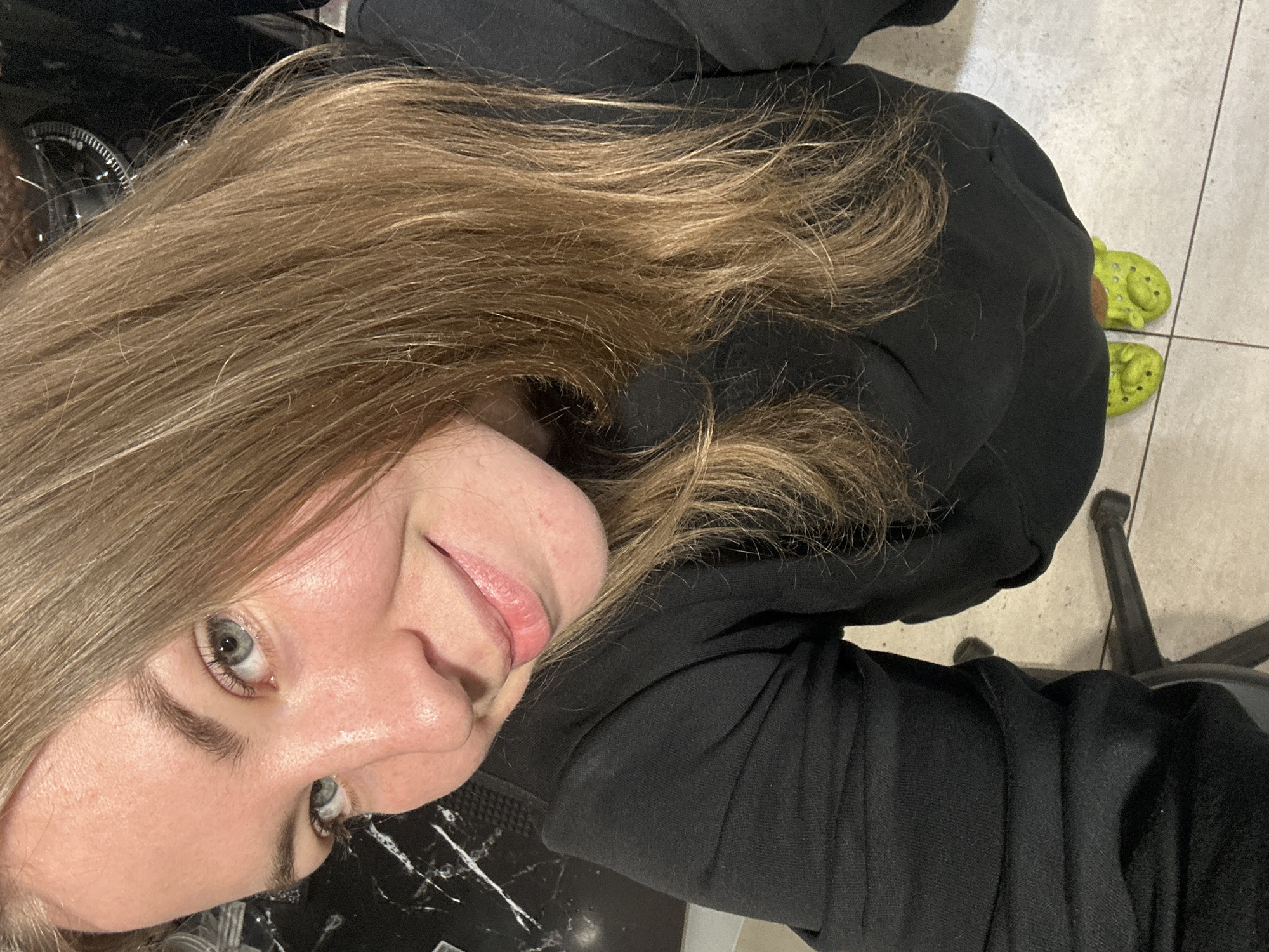 A high-angle selfie, slightly rotated, captures a person looking directly at the camera with a subtle, relaxed smile. The individual has long, wavy hair, a mix of light brown and blonde tones, which frames their face and extends towards the bottom of the frame. Their eyes are a clear blue-grey, and their lips have a natural pink hue. They are wearing a casual black long-sleeved top, possibly a hoodie or sweatshirt, which contrasts with their hair and skin.
The lower right portion of the image reveals the person's feet, clad in distinctive bright lime green Crocs. These Crocs are adorned with a character design, featuring what appears to be a cartoon face and ears, strongly resembling the character Shrek. The setting appears to be indoors, characterized by a light-colored tiled floor with visible grout lines, suggesting a casual environment. Dark, indistinct elements of furniture or equipment are visible in the background, including a black chair base. The lighting is uniform, indicating an indoor, possibly artificial, light source. No visible text is present in the image.