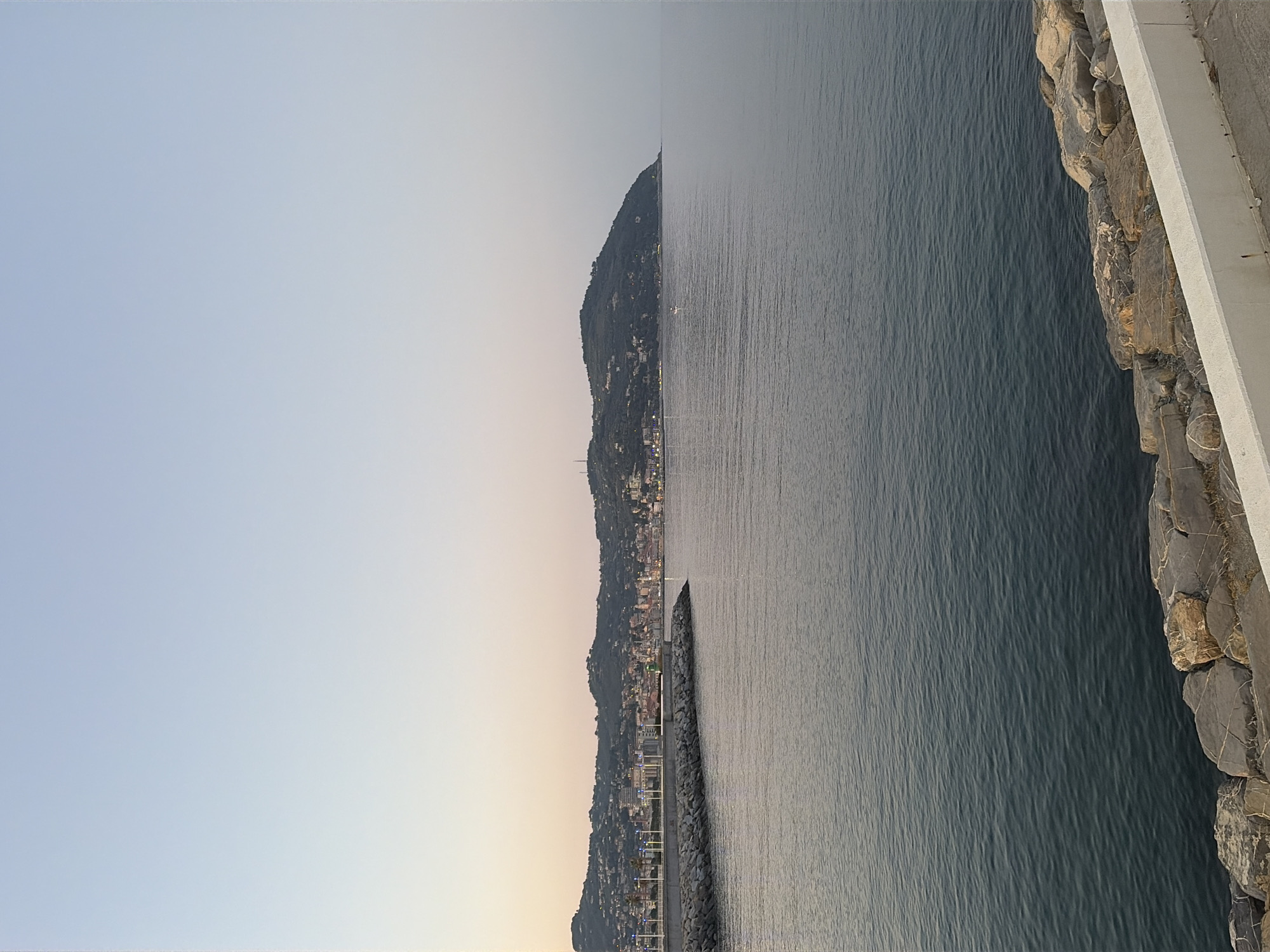 This is a landscape photograph taken from a waterfront promenade, looking out over a body of water, likely the Ligurian Sea. The image is oriented vertically, with the sky at the left and the sea and coastline to the right.
The left side of the image shows a pale, gradient sky, transitioning from a soft blue at the top to a warm, peachy hue near the horizon. This suggests it is either sunrise or sunset. The right side of the image is dominated by the deep blue water, with subtle ripples indicating a calm sea.
Along the distant coastline, a densely built town is visible nestled against a steep, tree-covered hillside. The buildings appear to be clustered together, with some lights twinkling, further supporting the idea of dawn or dusk. A breakwater, constructed of large stones, extends from the shoreline into the sea, creating a segmented line in the water. In the water, a small white boat with its lights on is visible.
In the foreground on the far right, there is a concrete walkway or promenade with a rocky embankment bordering the sea. The rocks are varied in size and color, with rough textures. The overall mood of the image is serene and tranquil, capturing a peaceful coastal scene at the cusp of day. There is no visible text in the image.