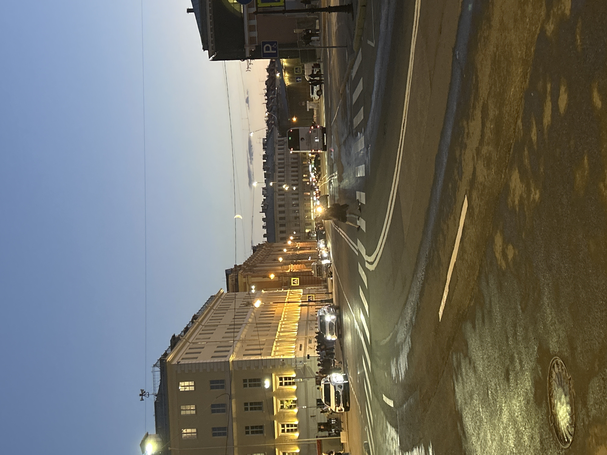 The image captures a street scene at dusk in an urban environment, possibly in St. Petersburg, Russia, given the architecture. The sky is a gradient of deep blue to lighter hues of orange and pink, indicating either sunrise or sunset. A crescent moon is faintly visible in the upper portion of the sky.
The foreground features a wet asphalt road with white lane markings. Puddles and reflections of streetlights are visible on the surface, suggesting recent rain or moisture. A manhole cover is present on the left side of the road.
Buildings line both sides of the street. The building on the left is a multi-story structure with a pale yellow facade, illuminated by numerous warm-toned lights emanating from its windows. The building on the right appears to be darker in color, with architectural details and lit windows. Streetlights, casting a warm glow, are spaced along the sidewalks.
Several vehicles are on the road. In the foreground, a white SUV is turning to the left. Further down the street, other cars, a bus, and a white van are visible. There are pedestrians on the sidewalks and one person, a woman in a dark coat, is crossing the street at a pedestrian crossing marked with white stripes.
The overall atmosphere is calm and urban, with the city transitioning from day to night. The presence of streetlights and illuminated buildings suggests the onset of evening activities.