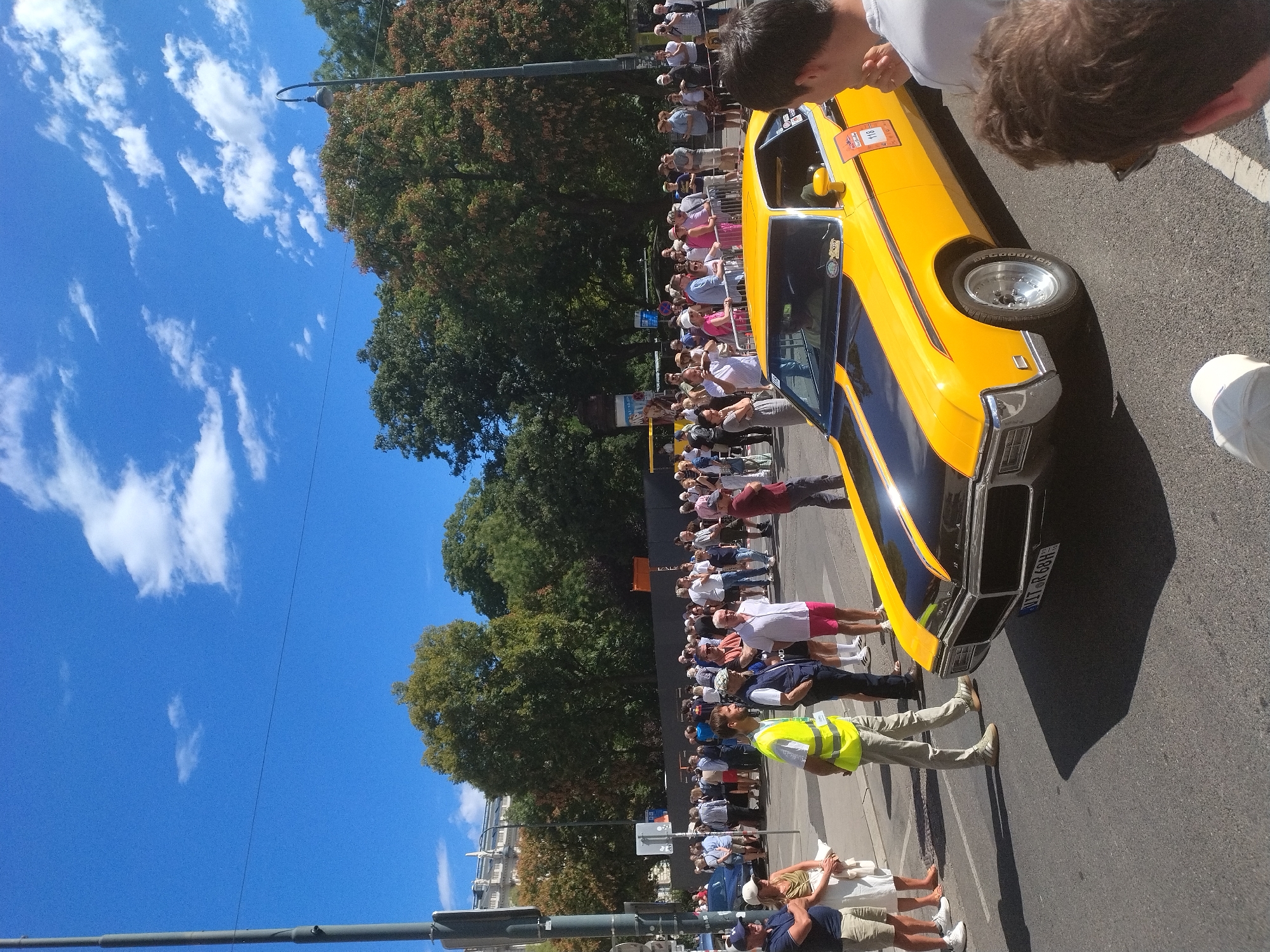 The image captures a street scene in Vienna, Austria, on a sunny day with a bright blue sky and scattered white clouds. A crowd of spectators lines a street, gathered behind a low metal fence. The focus of the scene is a vibrant yellow vintage car with black racing stripes, parked on the asphalt road. The car's license plate reads "WIR 68H". Several individuals are visible in the foreground and mid-ground. On the left, a man wearing a yellow safety vest and khaki pants stands facing the car. In the mid-ground, a group of people are casually dressed, some standing, others in conversation. The background features mature trees with lush green and some autumnal foliage, and a building with ornate architecture. A lamppost stands on the right side of the frame, and a tall pole with what appears to be traffic signals or signs is on the left. The overall atmosphere suggests an outdoor event or a parade.