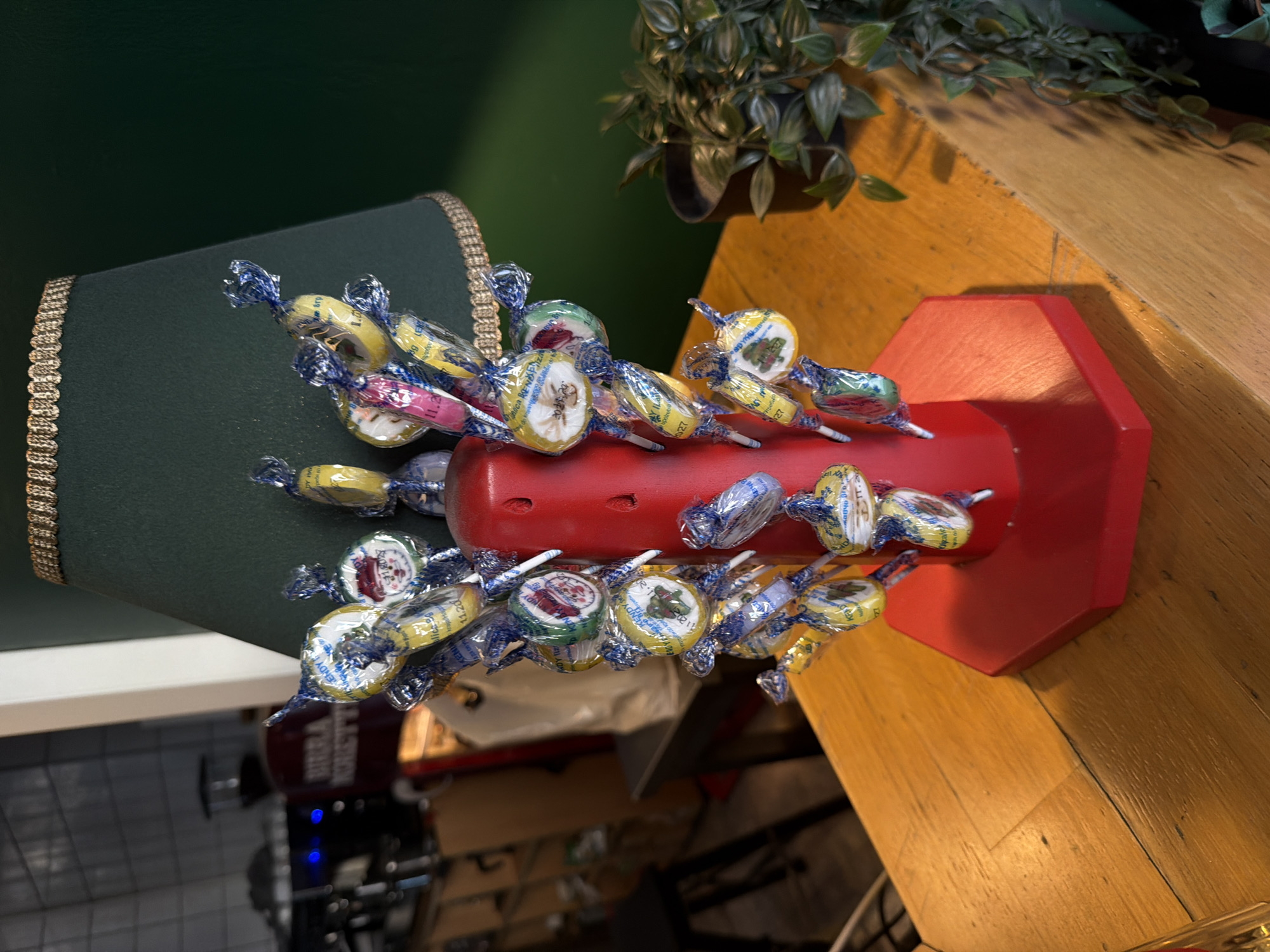 This is a close-up, eye-level, indoor, medium shot of a novelty item. The object is a red, hexagonal base topped with a cylindrical pillar adorned with numerous individually wrapped candies. The candies are arranged in a circular fashion around the pillar, with some appearing to be lollipops and others small, round sweets. The wrappers are colorful, featuring various designs including cartoon characters and holiday-themed images like Santa Claus and a Christmas tree. The pillar and base are a bright red, and the candies are attached by short sticks. Behind the candy display, a dark green lampshade with a gold trim is visible, casting a shadow on the background. To the right of the display, a potted plant with dark green, leafy foliage is situated on a wooden surface. The wooden surface appears to be a tabletop or counter, made of light-colored wood with visible grain. In the background, blurred elements suggest an interior setting, possibly a cafe or shop, with tiled walls and some equipment visible. There are no people present in the image, and no actions or interactions are depicted. The lighting is artificial, creating soft shadows. The scene is static and appears to be a still life arrangement. There is no information available to determine the location within the city of Belgrade, Serbia.