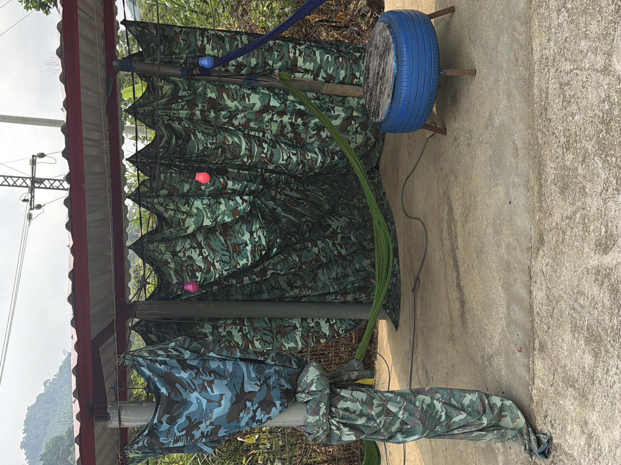 The image depicts a makeshift structure on a concrete surface. The structure is framed by metal poles and a corrugated metal roof with red trim. The sides are draped with camouflage fabric of two different patterns: one primarily green and brown, the other a blue-toned camouflage.
Suspended near the top of the structure are colored light bulbs: one blue, one pink, and one red. A green rope is draped around one of the poles and extends towards a blue painted tire mounted on a wooden frame, serving as a target.
The environment includes a backdrop of vegetation and a distant mountain range obscured by haze or fog. Electrical power lines are visible in the upper left portion of the frame. The exact location in Đà Nẵng, Vietnam, cannot be determined with certainty from the image alone.