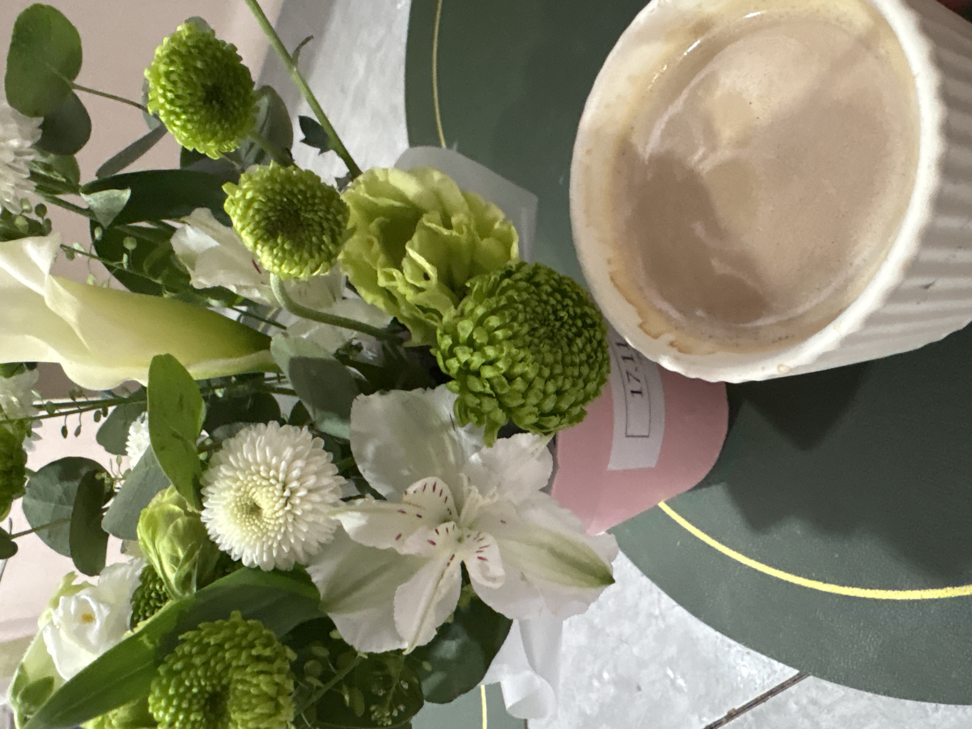A top-down view shows a bouquet of mixed flowers in white and green hues next to a mug of coffee on a green and gray surface. The flowers include white lilies, chrysanthemums, and a green carnation. A pink tag is visible near the base of the bouquet. The coffee mug is white and partially filled. The background features a surface with green and gray tones, hinting at a tabletop. There is no visible evidence to determine the location as Chaikovsky, Russia.