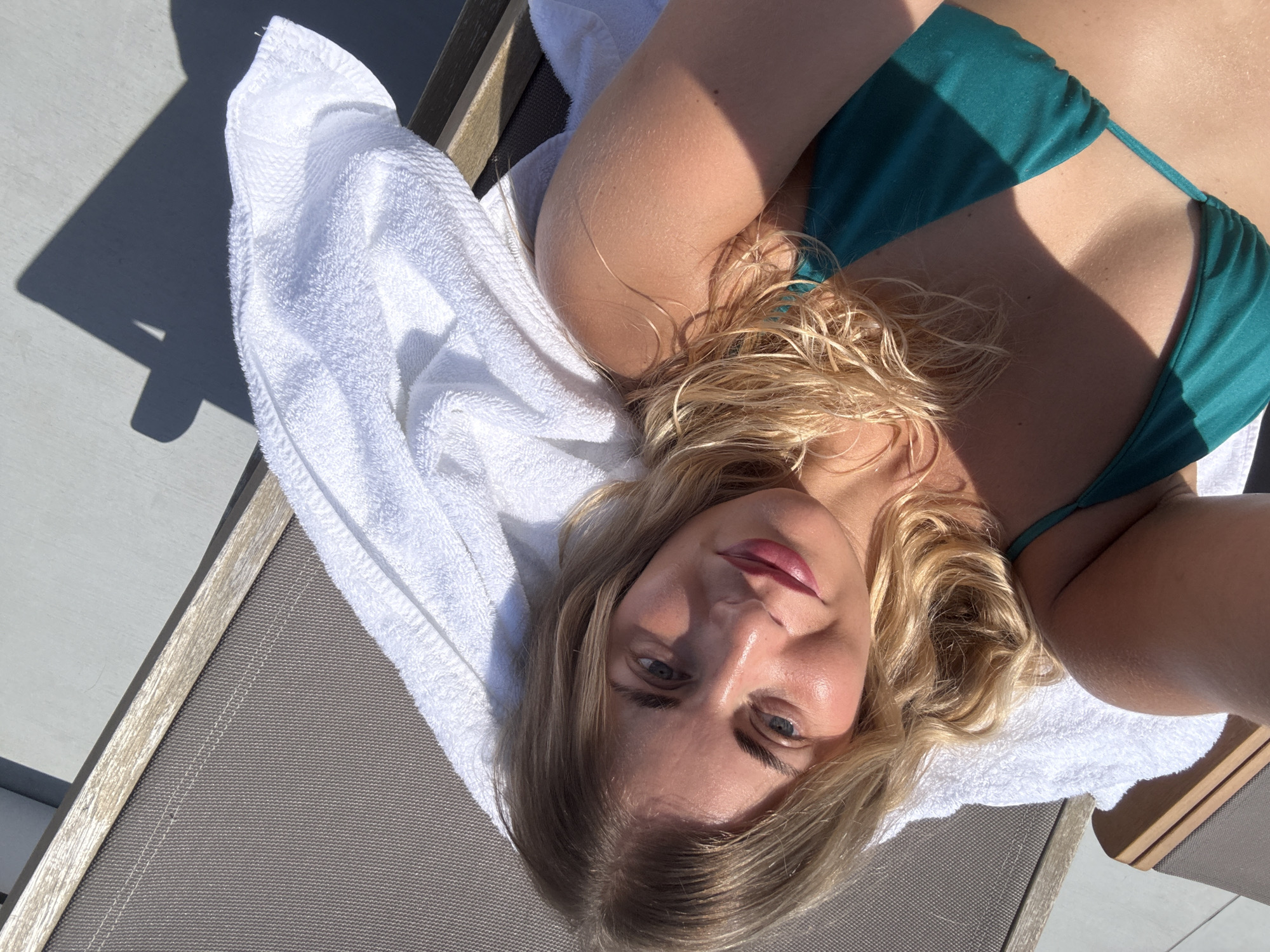 A blonde woman lies on a lounge chair outdoors, wearing a teal bikini top and a white towel. She is looking up at the camera and smiling. The sun is shining, casting a shadow of a chair on the ground behind her. The lounge chair has a grey mesh seat and wooden armrests.
