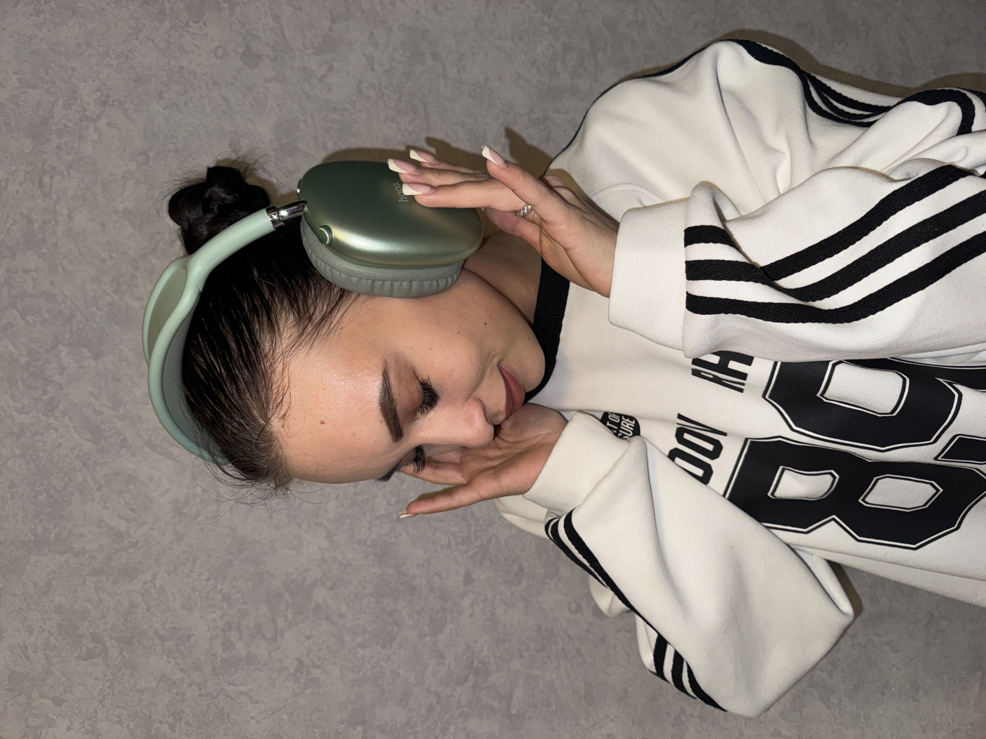 The media file features a young woman wearing over-ear headphones and a white sweatshirt in an indoor setting. The woman is positioned at an angle, smiling with her eyes slightly closed, suggesting contentment or enjoyment while listening to audio. Her dark hair is styled in a messy bun.
She is wearing green or mint-colored headphones; a visible logo on one ear cup identifies them as "hoco" brand. Her hands, adorned with manicured nails featuring white tips and a ring with small stones on her left ring finger, gently touch the headphones and rest near her chin.
Her white sweatshirt features distinctive black triple stripes on the sleeves, resembling an athletic style. Large black numbers "88" are prominent on the front of the sweatshirt, along with partial text that appears to read "OUT OF LEISURE" and "BODY RE". The background is a plain, textured gray surface, possibly a wall or floor. The lighting suggests an indoor environment.
The location context provided is Chaikovsky, Russia.