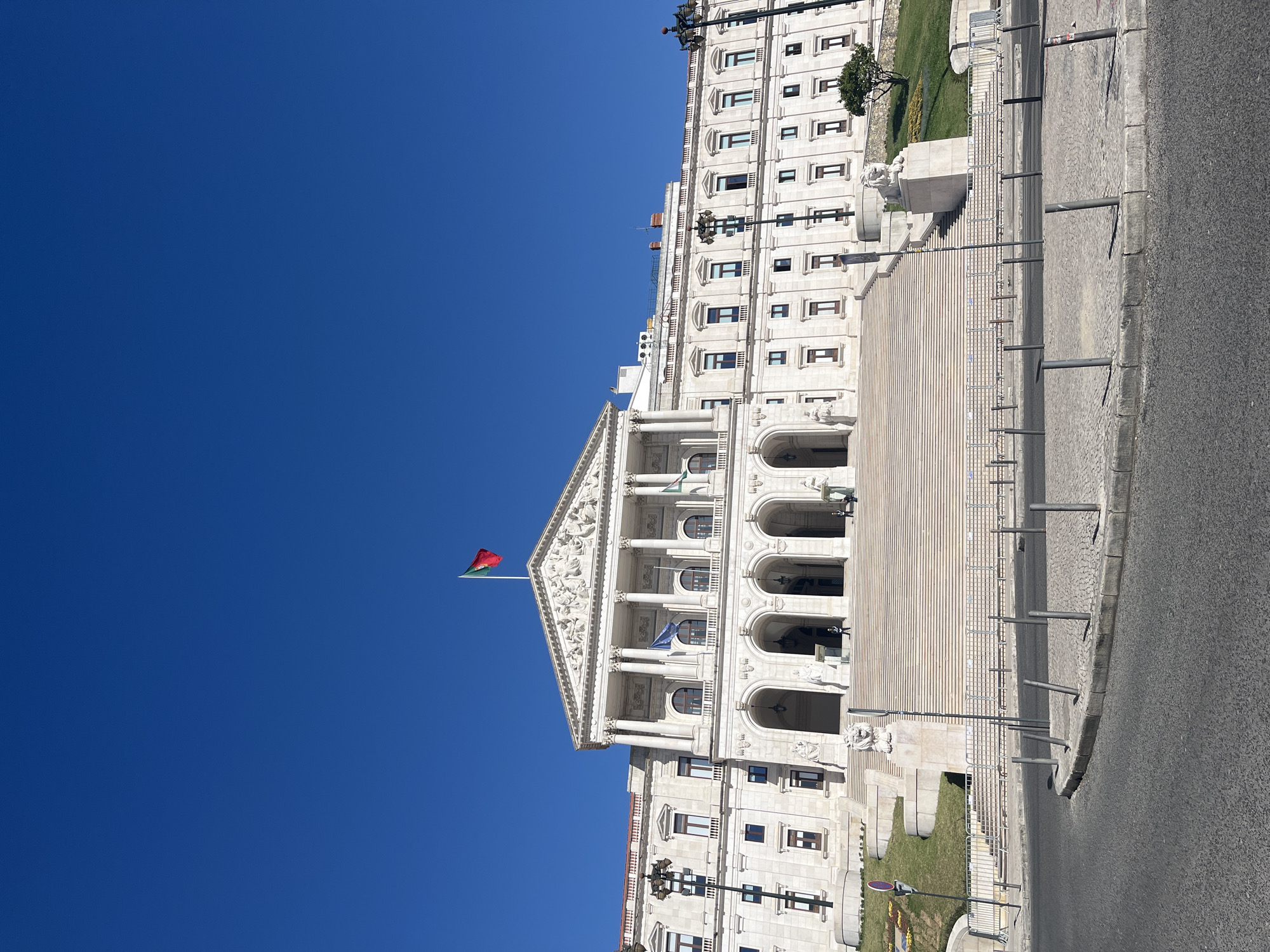 A daylight, eye-level shot shows the Assembly of the Republic building in Lisbon, Portugal. The camera is positioned at ground level on the street, angled upwards and slightly to the left, capturing the building's ornate facade and the clear, deep blue sky.
The Assembly building is white, with a classical design featuring a prominent pediment and a colonnaded front. Arches define the building’s entrance, and multiple windows are regularly spaced across its height. The Portuguese flag is visible atop the building, fluttering gently in the breeze. A European Union flag is also visible.
The building's steps lead down to the street level, where metal poles are situated at the curb. To the right of the steps, a small patch of green grass provides a contrast to the gray concrete steps and the asphalt street. No people are visible in the scene.