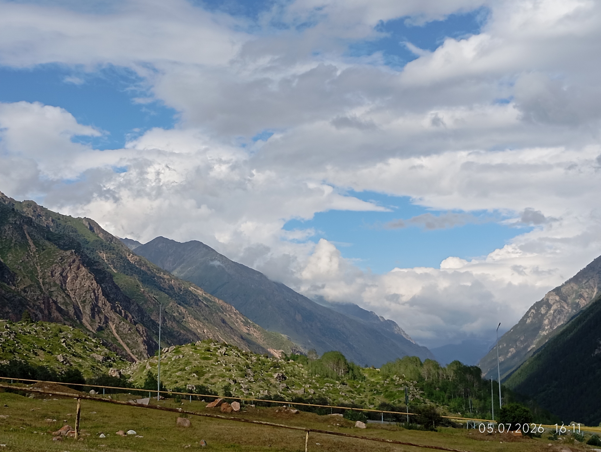 A wide, long shot captures a mountain valley under a cloudy sky. The foreground features a grassy field with scattered rocks and a yellow pipe running horizontally. A wooden fence with posts is visible in the bottom left. The middle ground is dominated by steep, rocky slopes covered in green vegetation and trees. A tall street lamp stands near the center of the frame. In the background, immense, dark mountain ranges stretch into the distance, partially obscured by clouds. The sky is a mix of blue patches and dramatic white and grey clouds. A timestamp in the bottom right corner reads "05.07.2026 16:11". The location is identified as Neitrino, Russia.