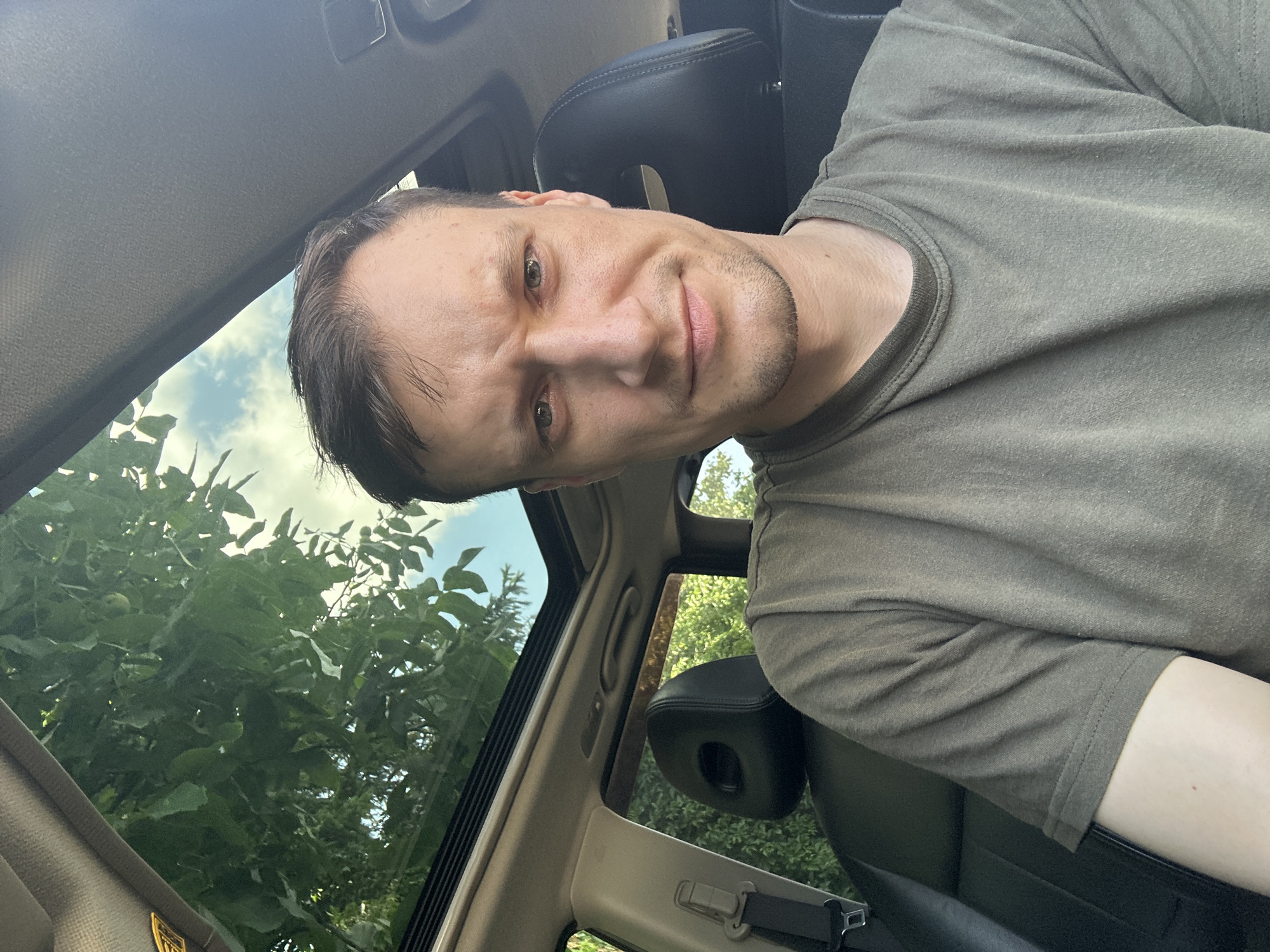 A medium shot shows a man in his early 30s with short brown hair and a light stubble sitting in the driver's seat of a car. He is wearing a green t-shirt. His facial expression is neutral, with a slight frown and his eyes are looking directly at the camera. The car's sunroof is open, revealing a clear blue sky with some clouds and lush green trees. The interior of the car is visible, including a black leather seat and part of the dashboard. The lighting suggests it is daytime, likely in the afternoon, with the sun casting shadows and highlights on the man's face and the car's interior. The background appears to be an outdoor setting, possibly a residential area or a park, given the presence of trees and foliage. The overall scene suggests a candid moment captured inside a vehicle, possibly while driving or stopped. The location of Shatava, Ukraine, is not visually confirmed within the image.