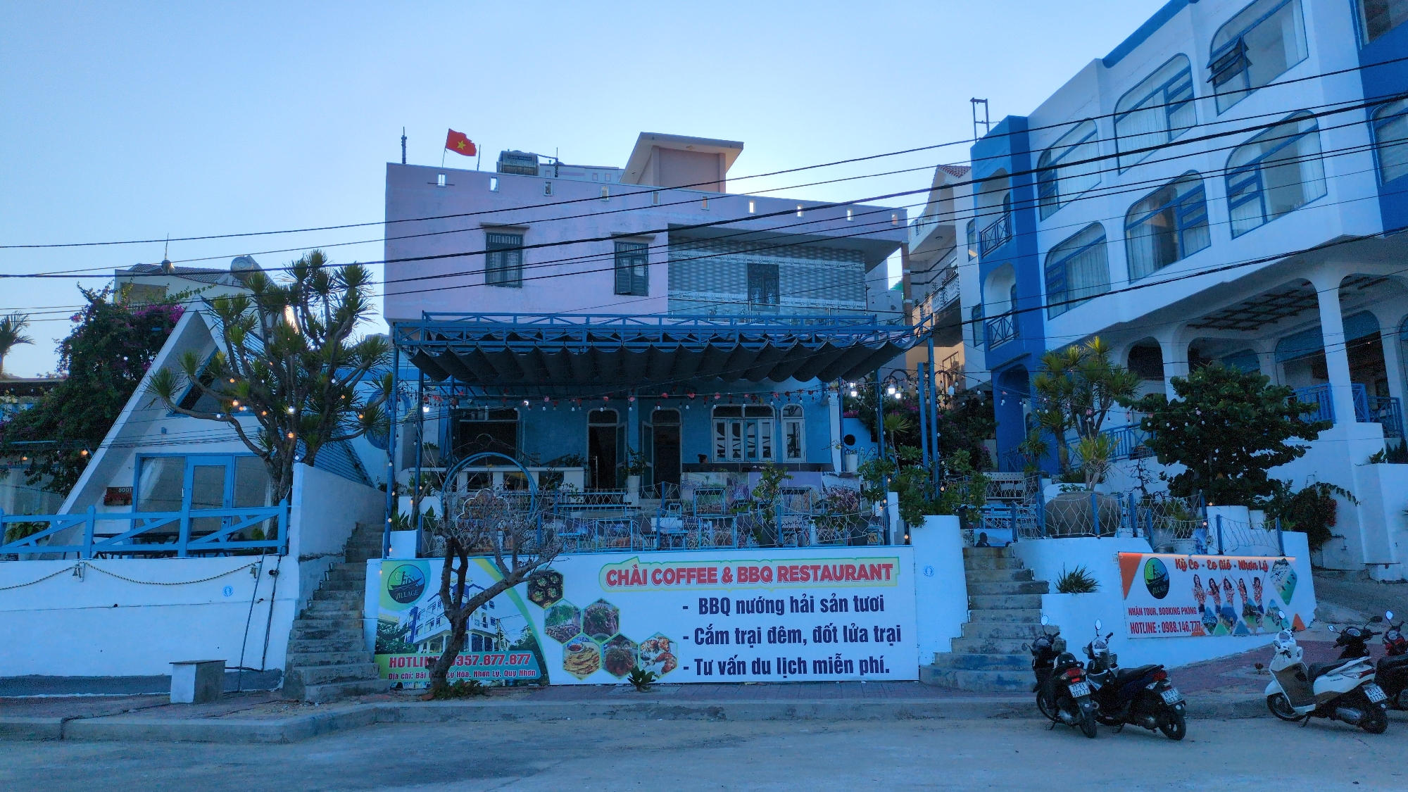 The image captures a street scene in East Quy Nhon Ward, Vietnam, under a clear, light blue sky. No individuals are present in the frame.
Three multi-story buildings are prominently featured. On the far left, a white, A-frame structure with blue trim includes a blue-framed glass door and a blue decorative fence, with the number "8001" visible near its base. A tree adorned with string lights stands in front of this building.
The central building, identified by a large white sign as "CHAI COFFEE & BBQ RESTAURANT," is a two-story structure. The lower level is blue, featuring multiple windows and an outdoor patio covered by a dark blue retractable canopy. The upper level is light pink with dark-framed windows. A red and yellow Vietnamese flag flies from the rooftop of this building. Multiple black electrical power lines horizontally cross the upper-mid section of the image, obscuring parts of the central and right buildings.
To the right, a multi-story building is predominantly white with blue accents, including arched windows, balconies, and support columns on its lower level.
In the foreground, a concrete sidewalk runs parallel to the buildings. A prominent white sign for "CHAI COFFEE & BBQ RESTAURANT" displays orange text, Vietnamese descriptions of services (e.g., "BBQ nướng hải sản tươi"), and hexagonal images of food and activities. To the right of this, a smaller orange and white sign advertises tours and bookings. Several motorbikes, including at least two dark-colored and one white scooter, are parked on the sidewalk near the right edge of the frame. Concrete steps lead from the sidewalk up to the establishments. Sparse vegetation, consisting of small trees and potted plants, is present around the building bases and patio areas.