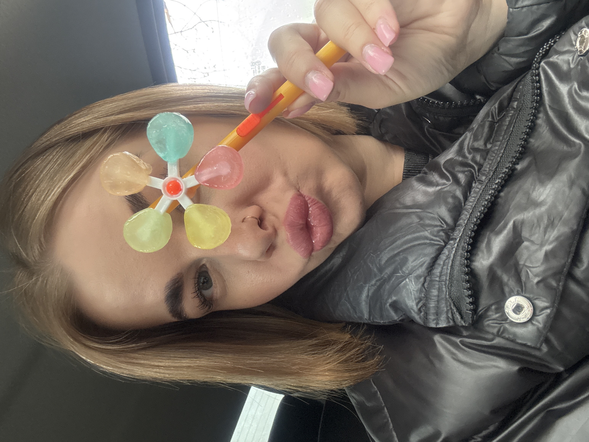 A woman poses for a selfie, holding a colorful lollipop-like object in front of her face. She wears a puffy black jacket and has light brown hair. Her nails are painted pink. The background is blurred, but suggests an interior space with a window. No information allows confirmation of the location in the city of Chaikovsky, Russia.
