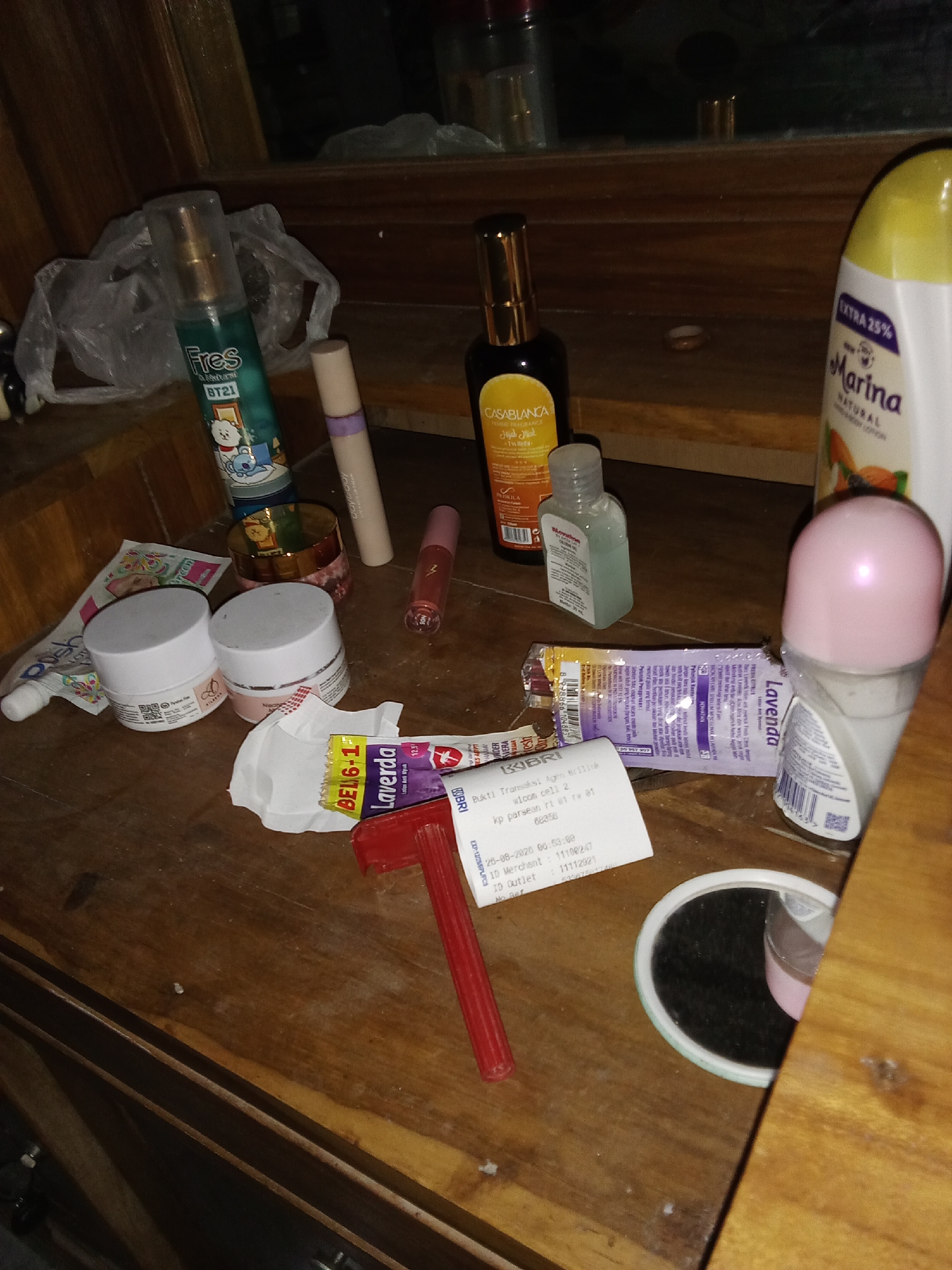 The image depicts a cluttered wooden surface, likely a tabletop or shelf, containing various personal care and cosmetic items. In the background, a window reflects a dimly lit indoor environment. Several items are scattered haphazardly, including a green spray bottle with a "FRES" label and a BT21 character, a light-colored tube, and a dark bottle with a gold cap labeled "CASABLANCA." There are also two white jars with pink lids, a small clear bottle of hand sanitizer, and a tube of toothpaste. A crumpled receipt and a red-handled razor are visible in the foreground, alongside a roll-on deodorant with a pink applicator. A yellow bottle of "Marina" body lotion is positioned on the right side of the frame. The scene is consistent with a personal vanity area or a collection of items left on a table in a residential setting. There is no indication of a specific location within the city of Selowogo, Indonesia.