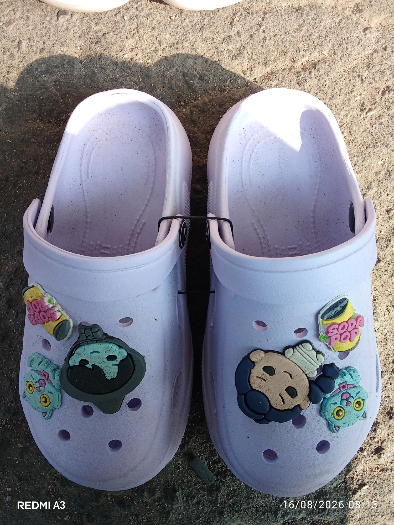 A pair of light purple clogs, each adorned with colorful Jibbitz charms, rests on a rough, textured surface, possibly concrete or dirt. The clogs are positioned side-by-side, with their openings facing upwards.
The left clog features three Jibbitz: a yellow soda can with red lettering that reads "SODA POP," a dark gray and turquoise cartoon character with a skull-like face, and a light blue cat-like creature with yellow eyes.
The right clog also has three Jibbitz: a yellow soda can with red lettering that reads "SODA POP," a cartoon character with brown skin and dark hair wearing a white hat, and a light blue cat-like creature with yellow eyes, similar to the one on the left clog.
A thin black strap extends across the top opening of each clog. The ground beneath the clogs is uneven and appears to be a mixture of sand and pebbles. Shadows are cast on the ground, suggesting the presence of sunlight. The lower left corner of the image displays "REDMI A3" and the lower right corner shows the date and time "16/08/2026 08:13". This scene is not located in Cemani, Indonesia.