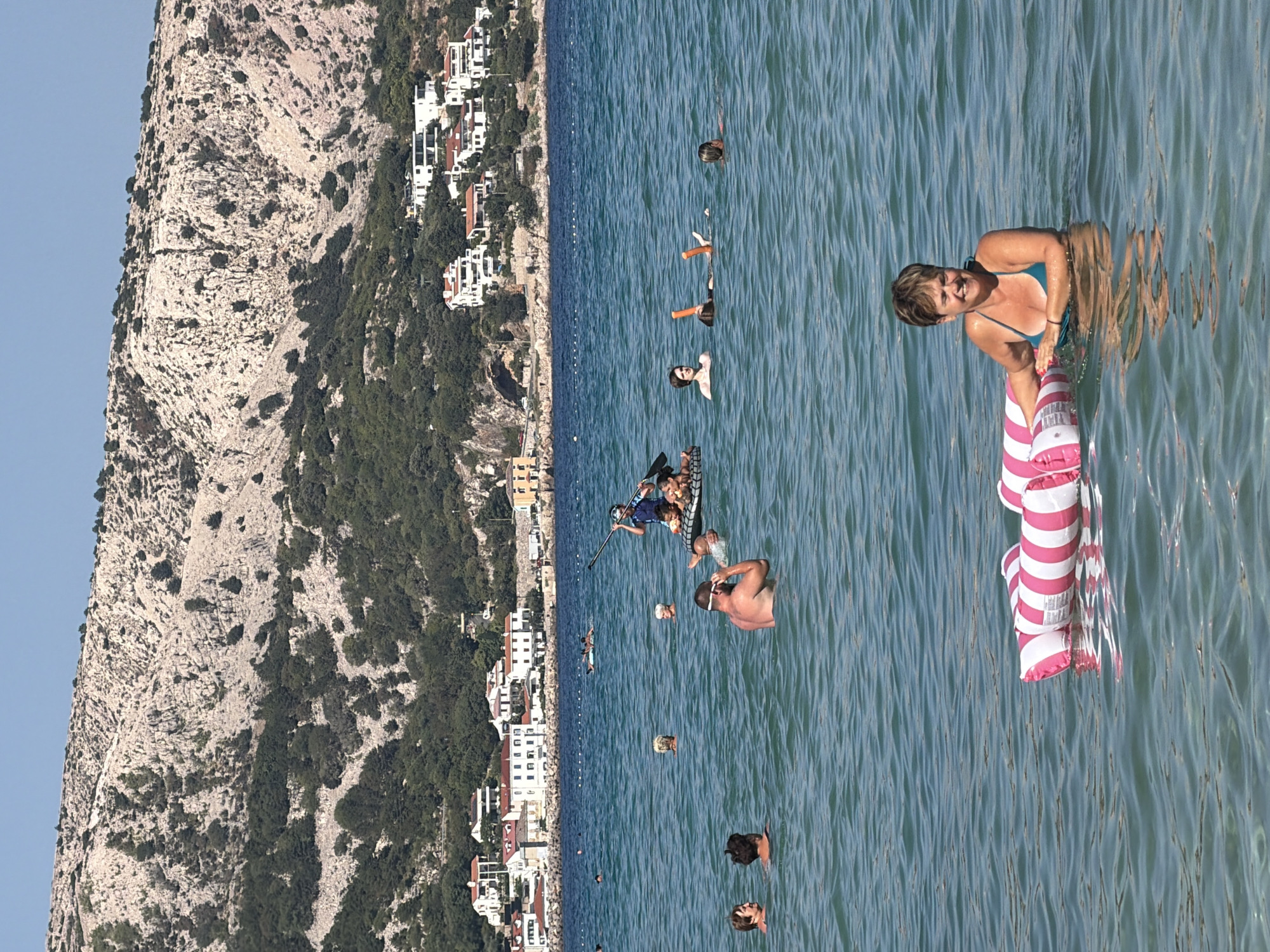 A group of people are enjoying a sunny day at the beach in Općina Baška, Croatia. The water is clear and blue, with gentle waves. In the foreground, a woman with short brown hair is floating on a pink and white striped inflatable mattress, smiling and looking to her right. She is wearing a turquoise bikini. In the background, other people are swimming and wading in the water. One person is on a paddleboard, paddling with a long oar. Further in the distance, a line of white buildings with red tile roofs are nestled against a rocky, tree-covered hillside. The sky is clear and blue.