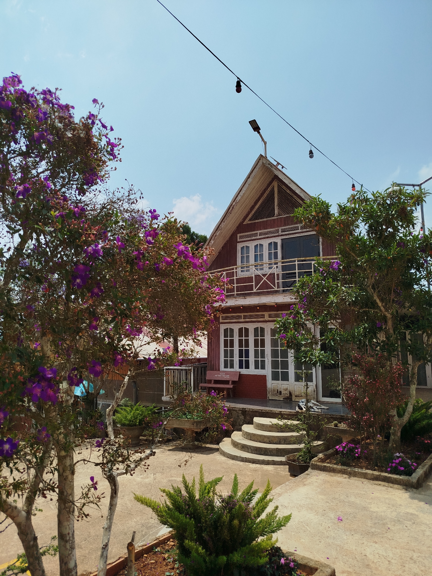 The image depicts a two-story house in Đa Thọ, Vietnam, surrounded by lush vegetation. The house is constructed of wood and painted a reddish-brown color. It features a gabled roof with a decorative triangular element, white-framed windows, and a balcony on the upper level. A string of lights hangs across the top of the image, with two bulbs lit.
The house is approached by a set of curved concrete steps leading to a porch. Trees with purple flowers frame the left side of the image, while green foliage is present on the right side. The ground is paved with concrete, and there are various potted plants and greenery. The sky is blue with some clouds visible.