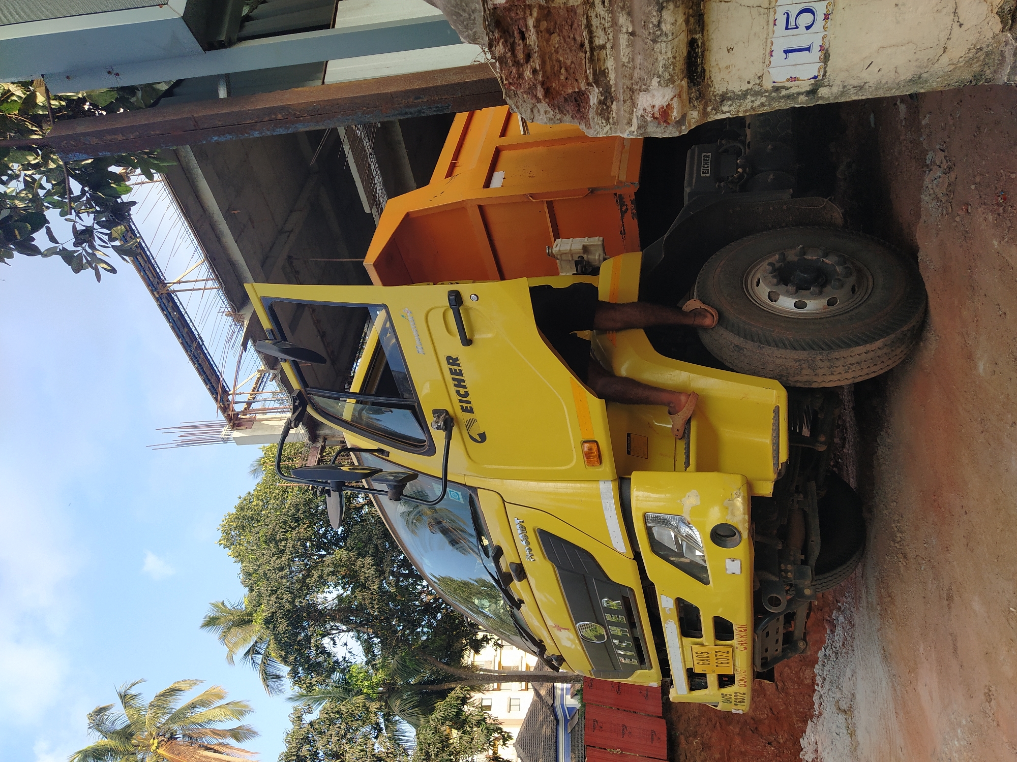 A yellow Eicher Pro 6019T truck is parked on a partially paved surface in a narrow space between a building and what appears to be a construction site. The building has a number '15' displayed on its exterior wall. A person's legs and feet are visible, seemingly stepping down from the truck's cab. The surroundings include trees, some showing the bright sky behind them. The location is Mapusa, India, as indicated by the license plate. The background includes building materials, possibly related to ongoing construction activities.