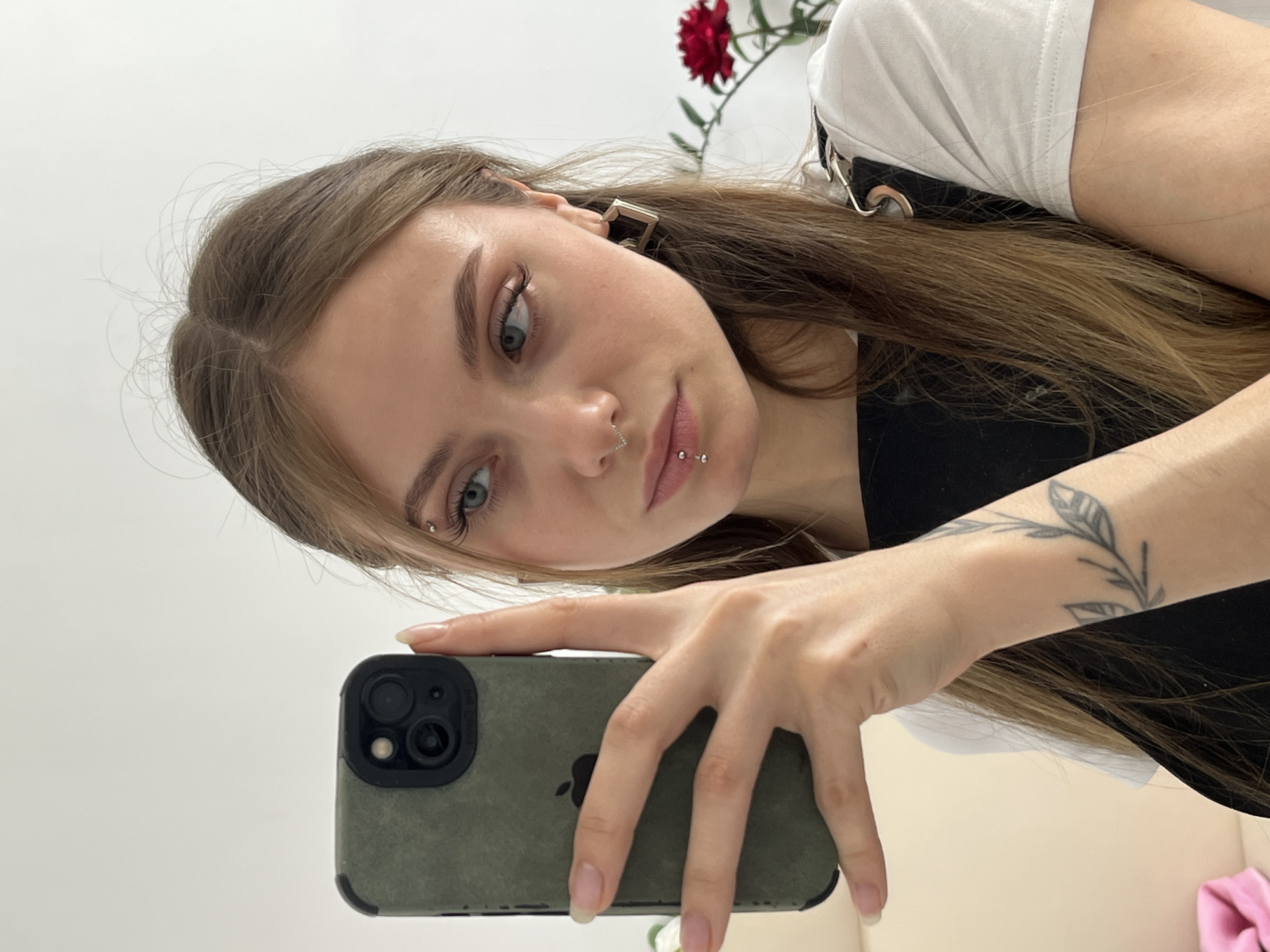 A woman is taking a selfie indoors with a smartphone. The phone has a green case. She has long brown hair and facial piercings in her nose, lip, and eyebrow. She is wearing a white shirt under a black outfit. A flower is visible in the upper right corner of the frame. The location is not discernable from the image.