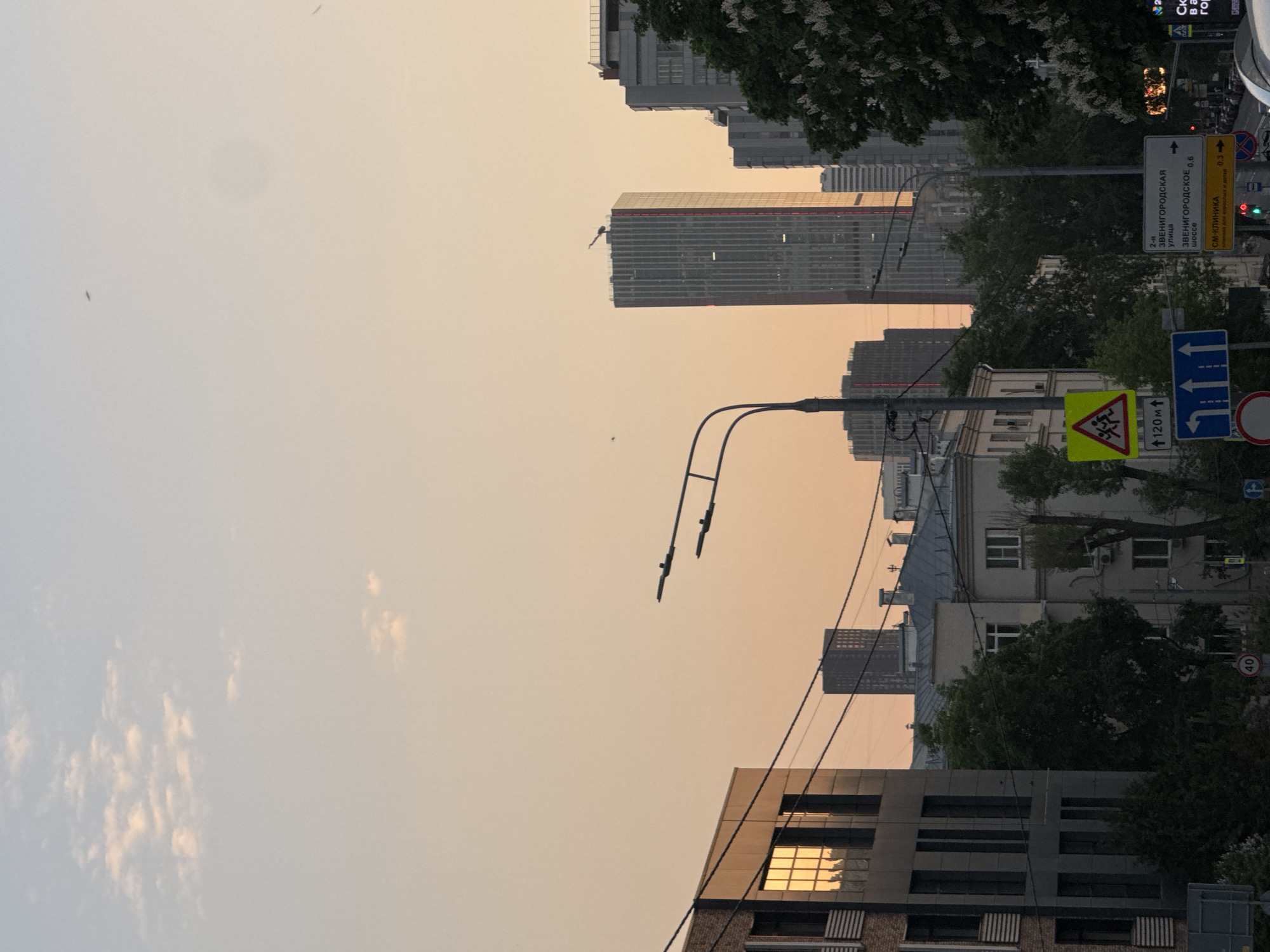 Here is a description of the image:
The image depicts a street scene in Moscow, Russia, recognizable by the street signs written in Russian and the architecture. The photo is taken at a low angle, looking up towards buildings, including tall modern skyscrapers, residential buildings, and a lamp post. Street signs, including those indicating directions to Zvenigorodskaya street and signs for a clinic, are prominent. There are trees and some greenery visible. The sky is a hazy orange color, likely due to smog or the time of day (sunset or sunrise). There are no people visible in the frame. The overall scene is an urban environment.