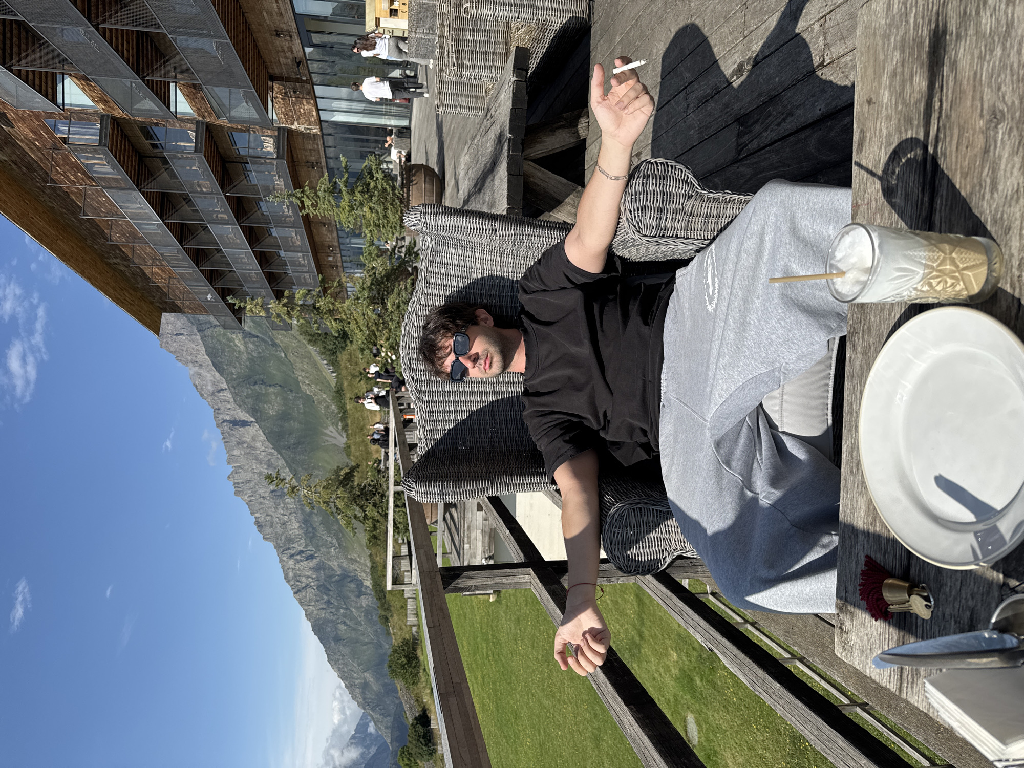 A man wearing sunglasses, a black t-shirt, and grey sweatpants sits in a wicker chair on a wooden deck in Stepantsminda, Georgia. He holds a lit cigarette in his right hand and has his arms outstretched. A drink with a frothy top sits on a wooden table in front of him, next to a white plate and a set of keys with a red tassel. In the background, a large wooden building with balconies and a mountain range loom under a clear blue sky. Other people are visible in the distance, some walking and others sitting at tables. The deck overlooks a grassy area and a wooden railing.