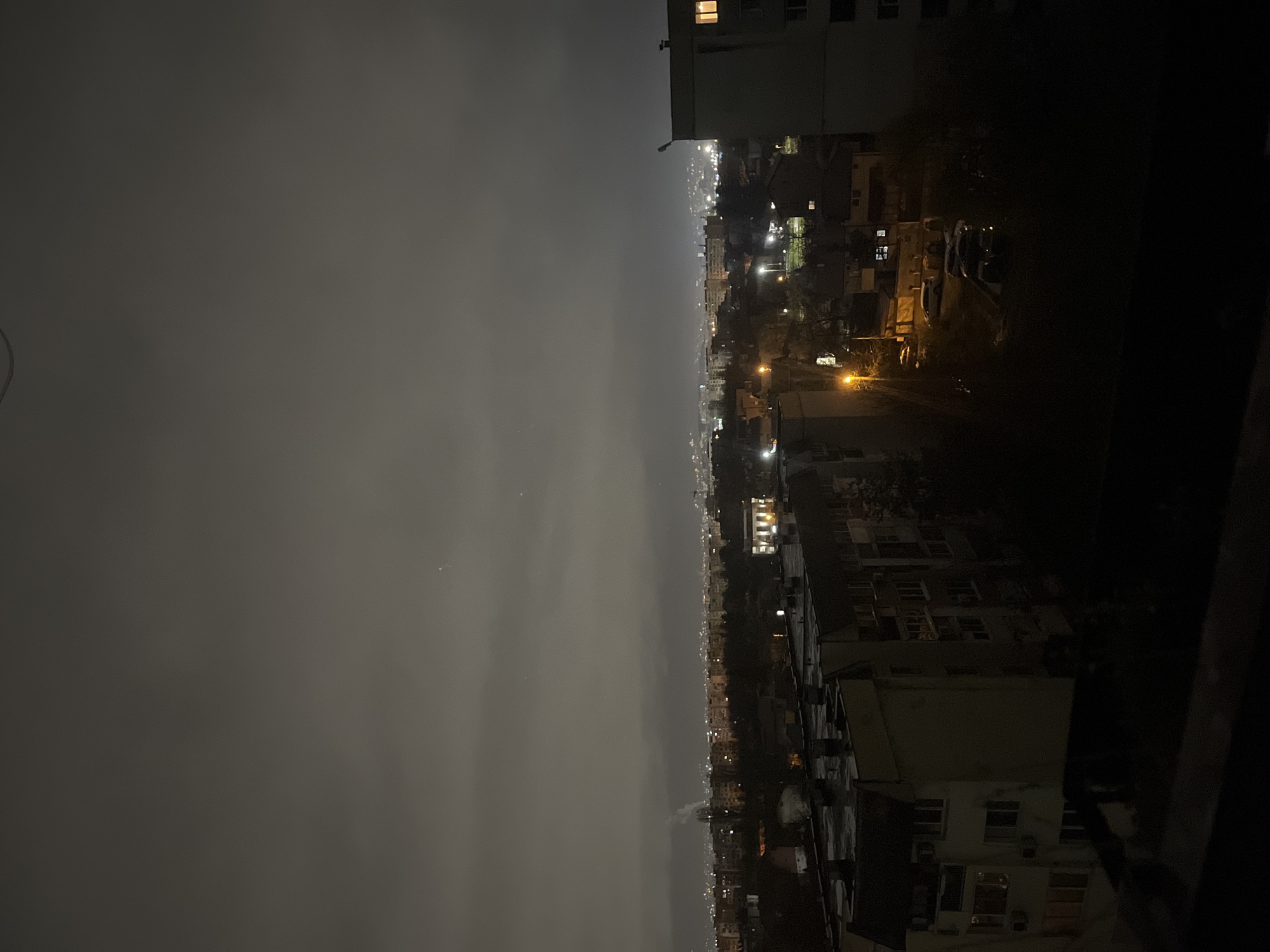 The image shows an urban night scene in Sofia, Bulgaria. The view is from an elevated position, likely a high-rise building, facing a cityscape. The sky is heavily overcast, creating a dim ambient light across the scene. In the distance, faint city lights are visible, suggesting a densely populated area. Closer to the viewer, there are residential buildings, some illuminated by streetlights or interior lights. The buildings vary in height and style, typical of Sofia's mixed architectural landscape. No people are visible.
