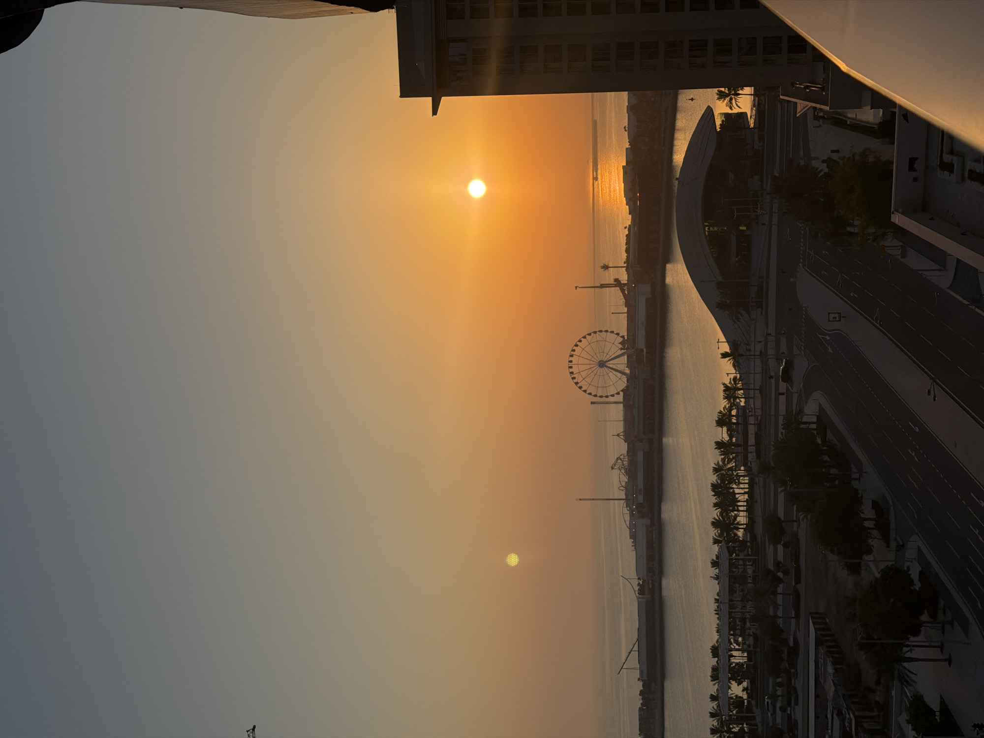 The image depicts a sunset over a coastal city. The sun, a bright orb with a visible patterned surface, is partially obscured by atmospheric haze, casting a warm orange glow across the sky and reflecting on the calm water. To the right, a tall, modern building with many windows dominates the foreground, its facade partially illuminated by the setting sun. In the middle ground, across a body of water, a Ferris wheel stands as a prominent landmark, part of an entertainment district or boardwalk. Various structures, including what appears to be a roller coaster and other amusement park rides, are visible along the waterfront, silhouetted against the bright sky. Palm trees line the waterfront and are also scattered throughout the urban landscape in the foreground, where roads and buildings suggest a developed city environment. The overall scene is one of urban development meeting the natural beauty of a coastal sunset.