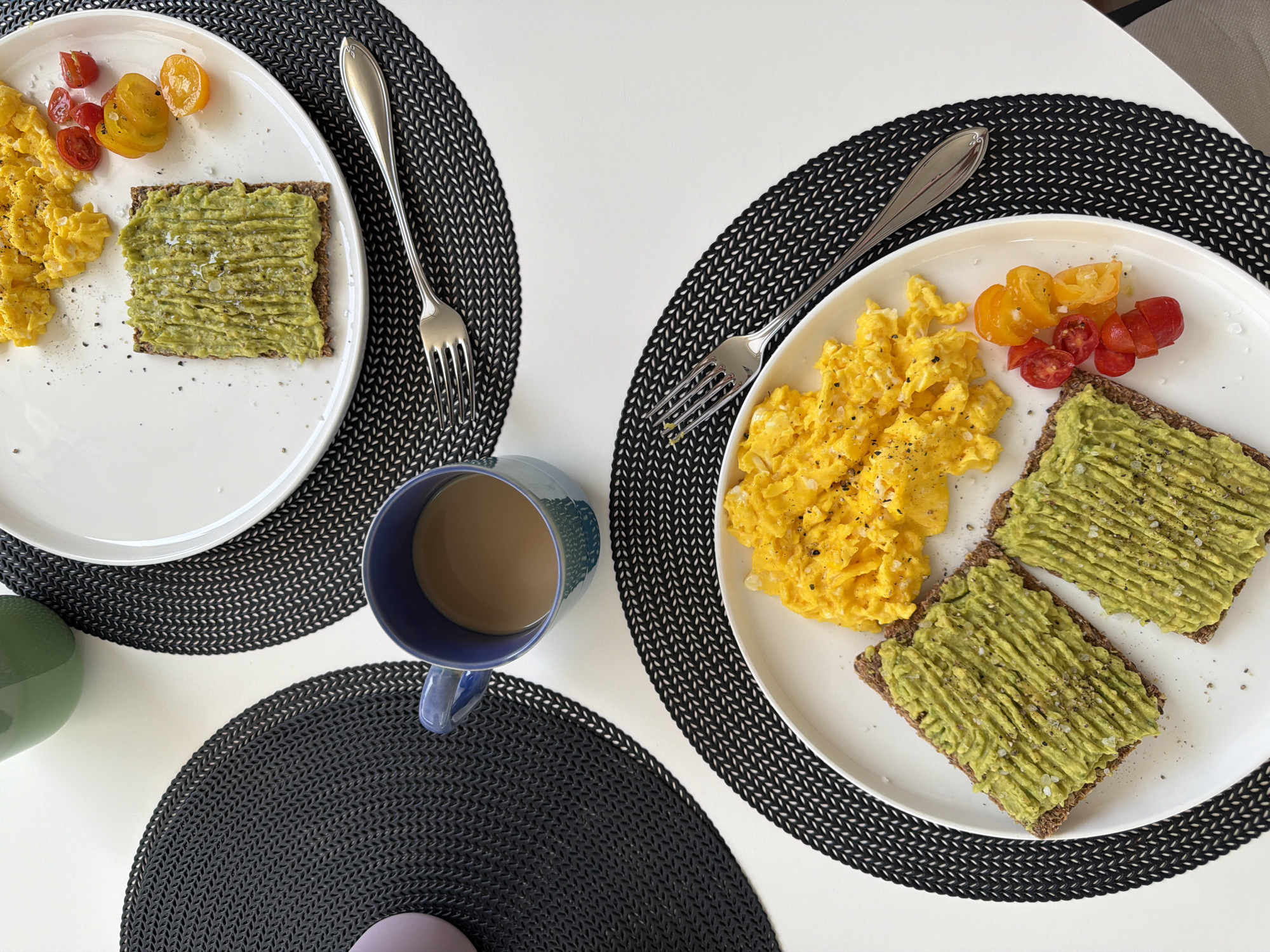 A high-angle, medium shot captures two plates of breakfast on a white tabletop, each adorned with a black placemat. The plate on the left displays two pieces of toast topped with mashed avocado and sprinkled with salt and pepper, alongside a portion of scrambled eggs and halved cherry tomatoes. The plate on the right features one piece of avocado toast, scrambled eggs, and cherry tomatoes. A blue mug filled with coffee sits between the plates on a placemat. The background is uncluttered, with hints of a purple object and a green vase visible. There are no people present in the image.