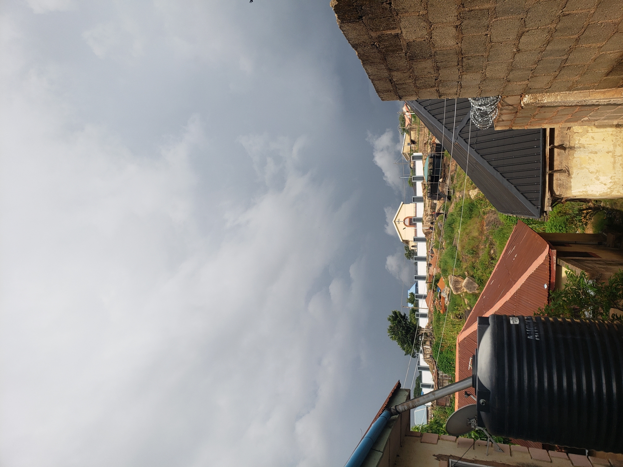 The image captures a view of the sky and rooftops in Jos, Nigeria. The sky is overcast with dark, dramatic clouds, suggesting an impending storm. In the foreground, a black water tank is visible on the left, attached to a building with a blue gutter and a rust-colored corrugated metal roof. To the right, a building constructed of rough, brown bricks occupies the right side of the frame. A sloped, dark metal roof with barbed wire fencing runs along its top. In the middle ground, a white building with a cross on its roof, resembling a church, sits on a rocky hillside. Scattered houses and trees are also present on the undulating terrain. The overall impression is an urban or semi-urban environment in a hilly region.