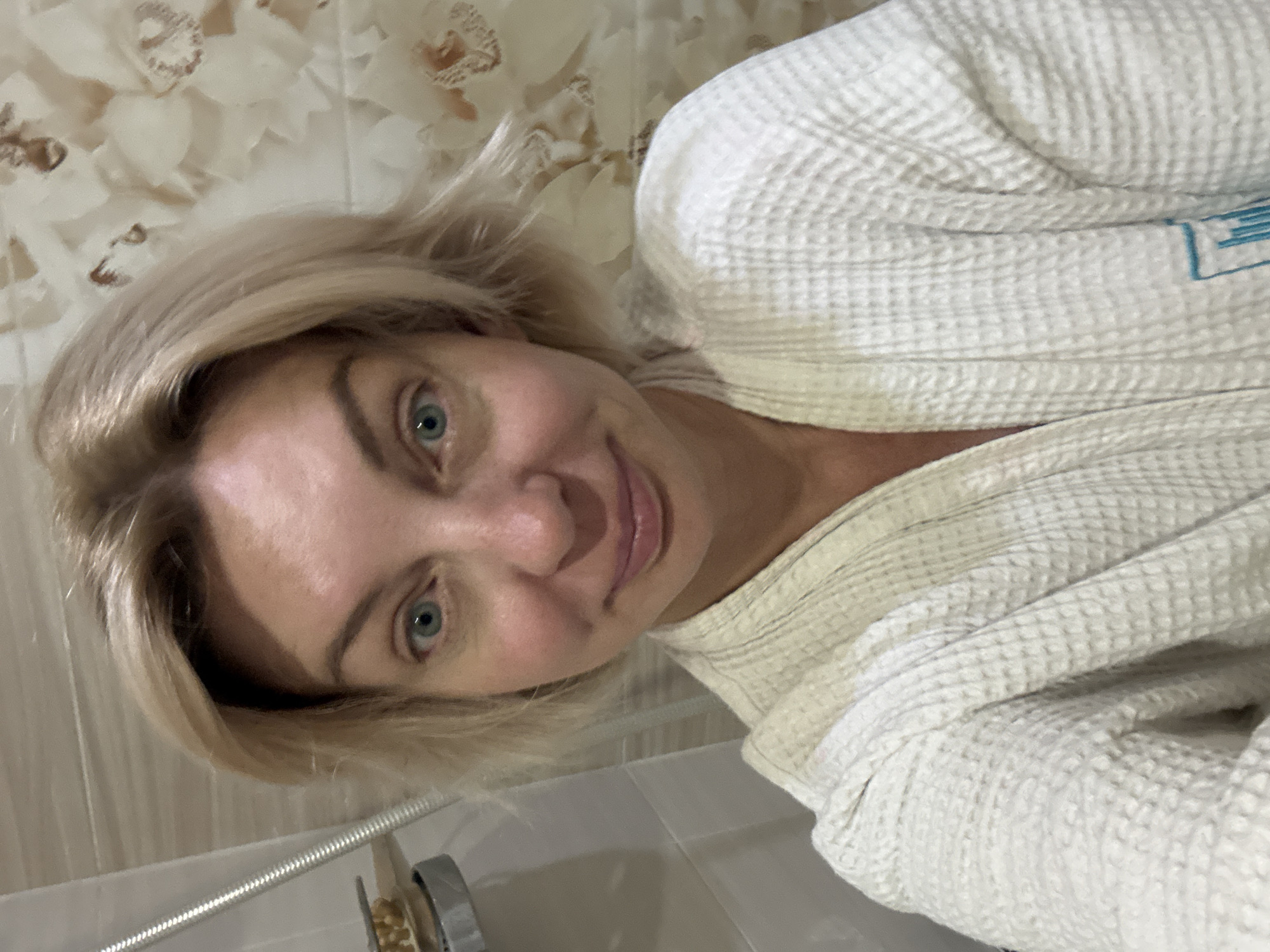 A close-up selfie of a woman with short blonde hair and blue eyes is shown. She is wearing a white waffle-knit bathrobe. The background appears to be a bathroom with tiled walls, one of which has a floral pattern. A shower head is visible to the left of the woman. The lighting is bright, suggesting it is daytime or well-lit interior. The woman is looking directly at the camera with a slight smile. There is no indication of the location being in Sochi, Russia.