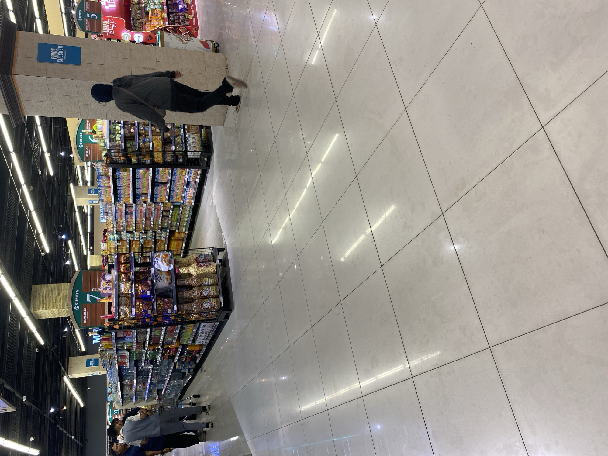 The image depicts the interior of a brightly lit supermarket, likely a branch of "SUZUYA" as indicated by signage visible on shelving units. The floor is tiled with large, light-colored square tiles, reflecting the overhead fluorescent lighting. Several aisles of merchandise are visible, stocked with various food and beverage products, including packaged snacks, drinks, and bottled goods.
In the foreground, the tiled floor occupies a significant portion of the frame, with the reflection of ceiling lights creating a pattern of bright streaks. To the left, a group of three individuals are walking, appearing to be customers. One person, wearing a blue shirt and dark pants, is partially visible. Next to them, another person in a grey uniform or vest and grey pants pushes a shopping cart. A third individual, also in dark clothing, is visible behind them.
Further into the supermarket, a person in a dark hoodie and dark pants walks away from the camera, towards the right side of the image. This individual is carrying a shoulder bag. They are walking past a large pillar constructed with stone-like materials.
Signage for "SUZUYA" is prominently displayed, including a large green sign with "BEVERAGE" and a number "7" beneath it. Other signs in English and potentially another language (likely Bahasa Indonesia, given the mention of Banda Aceh) are visible, such as "PRICE CHECKER". Red promotional displays are also present, featuring products like "TANGO" and "CHITO".
The overall environment suggests a typical supermarket setting with organized shelves, a clean and well-maintained floor, and ample lighting. The presence of the "SUZUYA" branding strongly suggests this location is within a supermarket operated by that chain. Based on the provided context of Banda Aceh, Indonesia, this scene is set within a commercial establishment in that city.