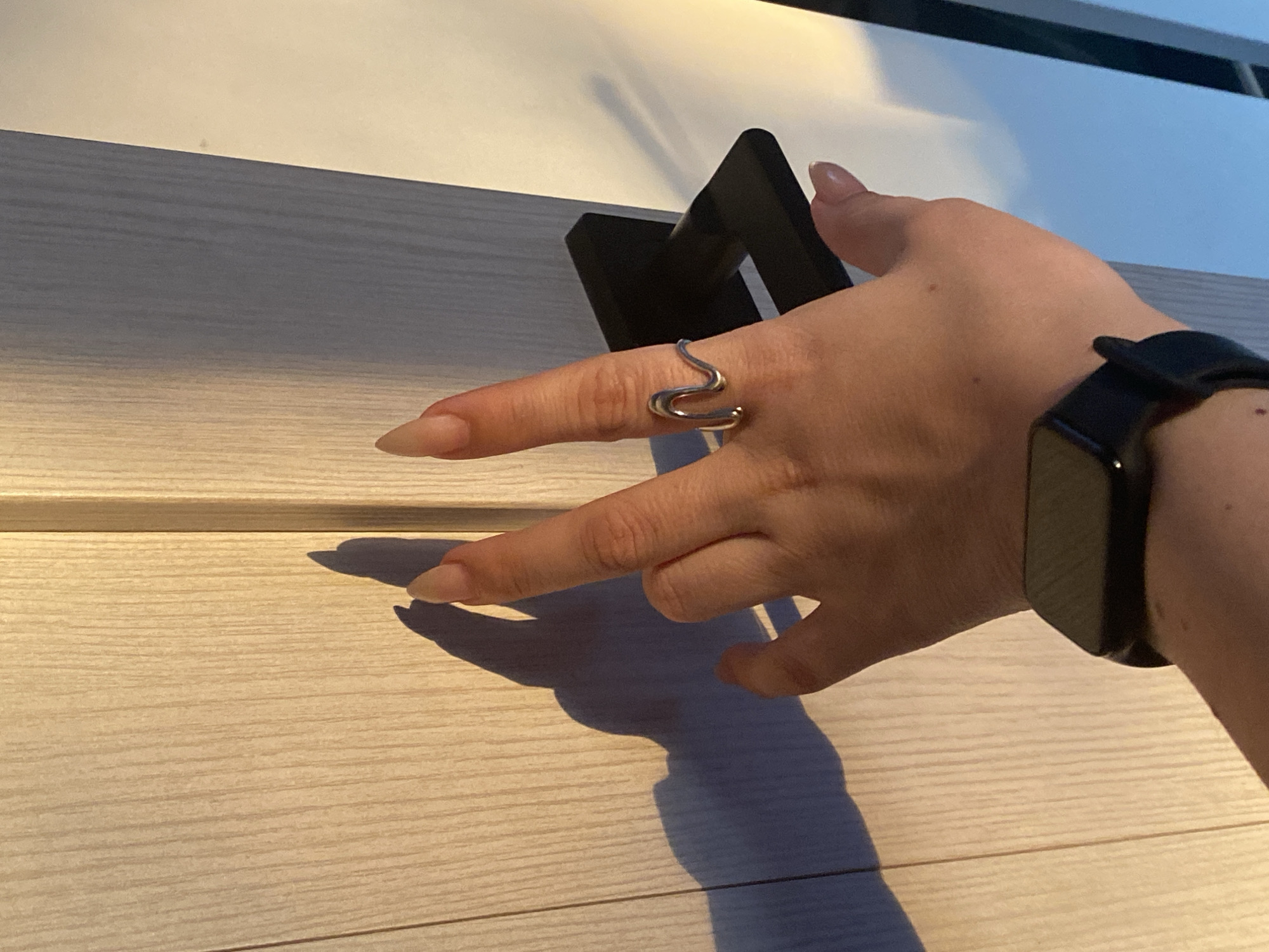 A fair-skinned hand with long, tan nails is holding a silver ring on the index finger. The ring has a wavy design. A black smartwatch is on the wrist. The background consists of a partially visible black door handle and a white, horizontally-patterned surface. The location is unknown, but the prompt suggests Chaikovsky, Russia, though there is no independent evidence to confirm this.