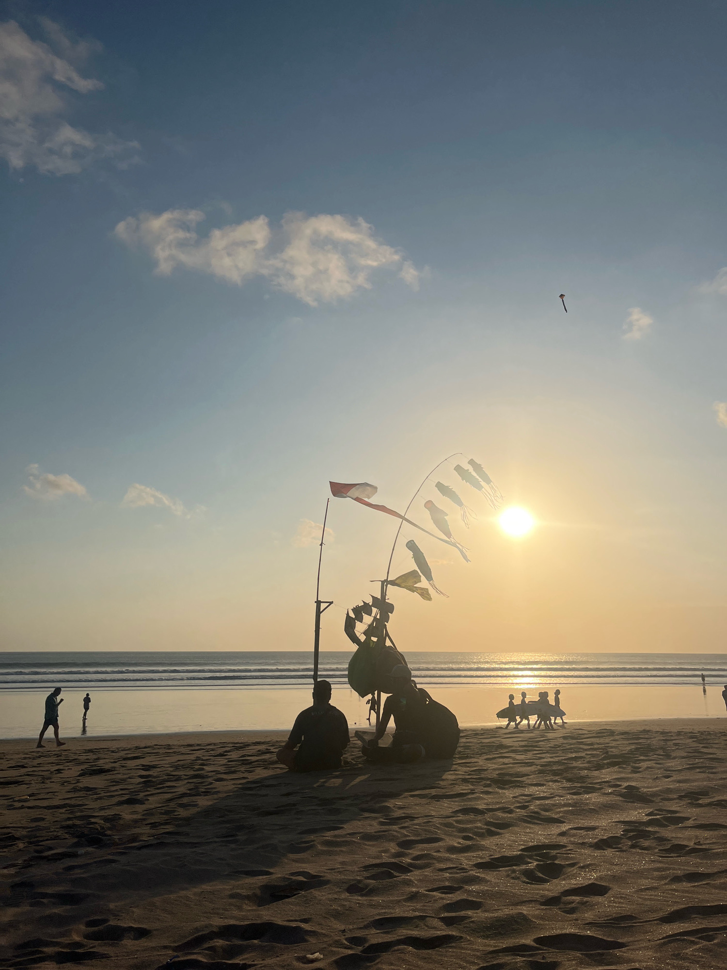 The image depicts a beach scene in Kuta, Indonesia, during sunset. The sun is low on the horizon, casting a warm golden glow across the water and sand. Several people are visible on the beach. In the foreground, a person is sitting cross-legged on the sand. To the right of them, a group of people, possibly surfers, are walking with surfboards. In the middle ground, a few more individuals are walking along the shore. In the background, the ocean stretches to the horizon with gentle waves. A kite is flying high in the sky. There are also decorative flags or banners on a pole near the center of the image, one of which appears to be the Indonesian flag. The sand in the foreground shows footprints and ripples. The overall atmosphere is peaceful and serene.