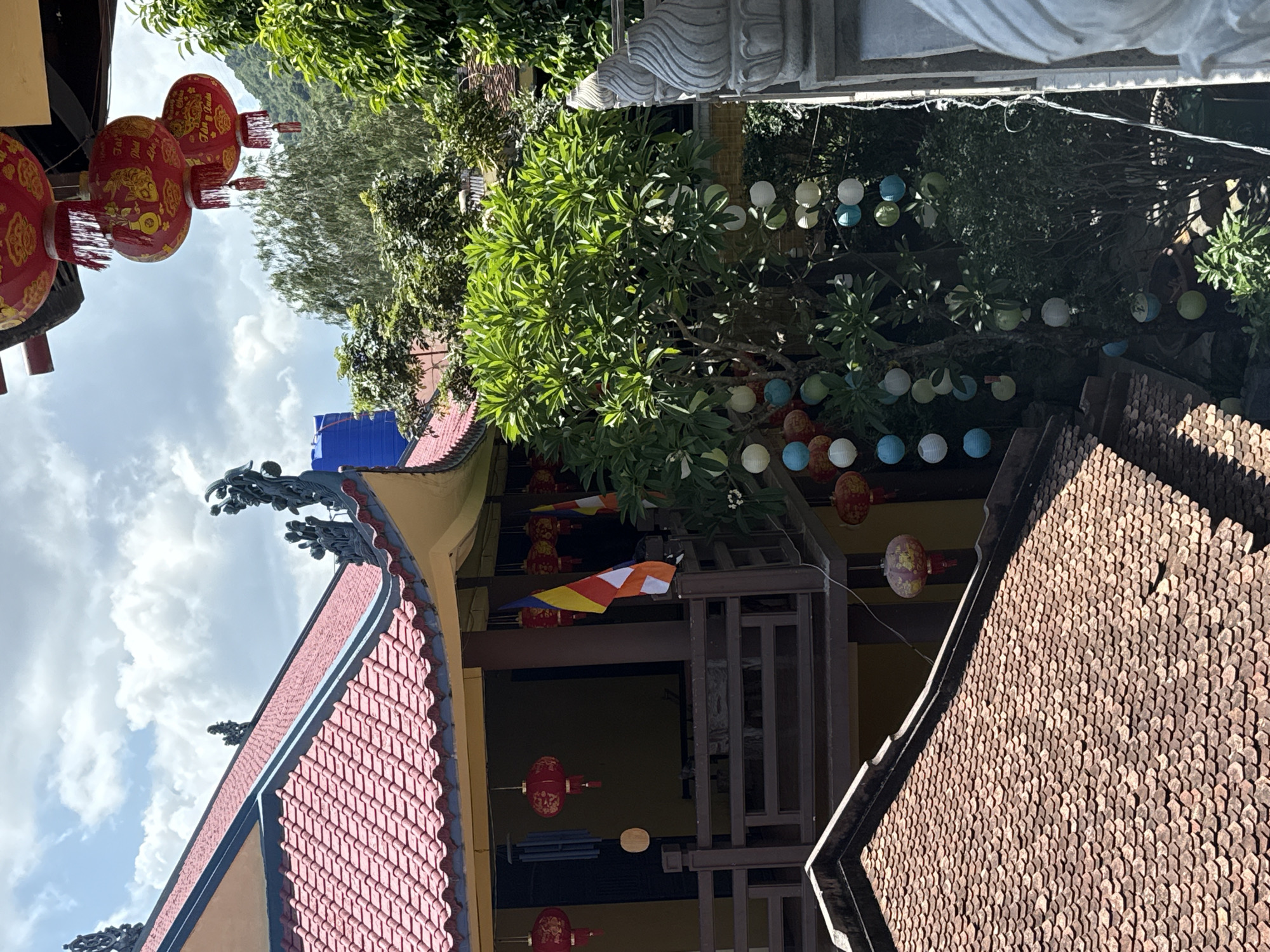 The image shows a building in Tay Nha Trang Ward, Vietnam. The building has a traditional Asian style with a red tiled roof and decorative eaves. There are red lanterns hanging from the roof and trees. The sky is cloudy. There are also colorful spherical lanterns hanging from the trees.