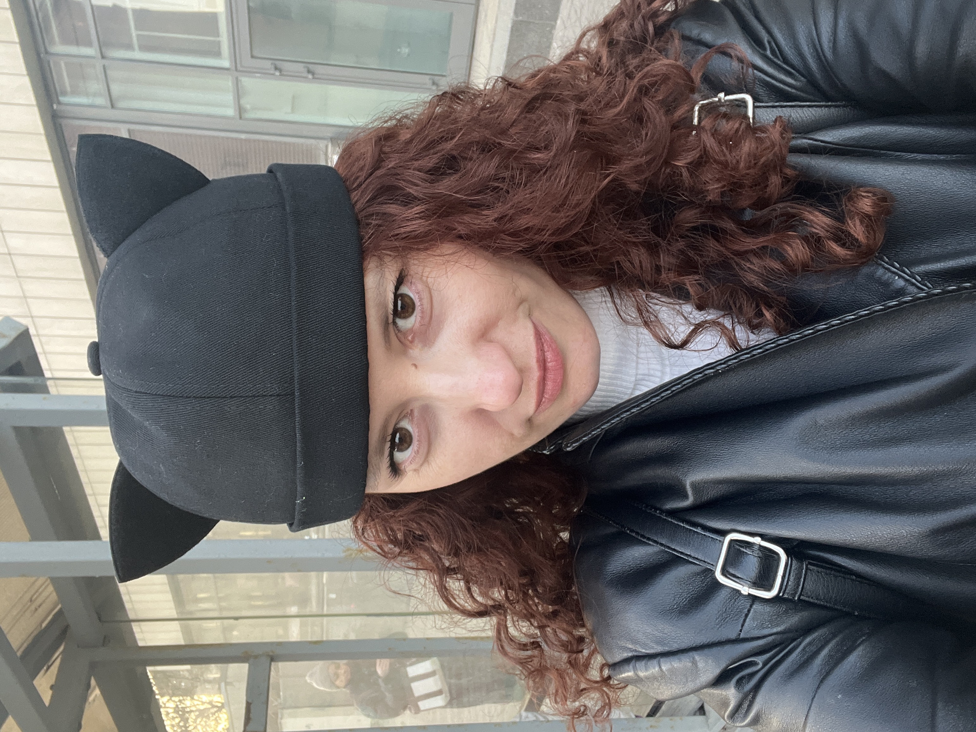 A woman with auburn curly hair and brown eyes, wearing a black cat-eared hat and a black leather jacket with a silver buckle, looks at the camera. She is outdoors. The background includes metal railings and a building with windows, suggesting an urban setting. The presence of other people blurred and reflected in the windows, combined with the architectural style, may indicate the image was taken in Saint Petersburg, Russia, but this cannot be determined with certainty.