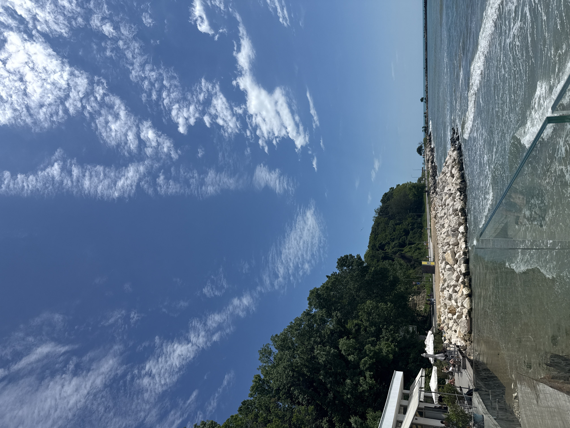 This image, set in Varna, Bulgaria, captures a bright and sunny day along a picturesque coastline. The upper portion of the frame is dominated by a clear blue sky, adorned with scattered, wispy white clouds forming distinct, elongated streaks. To the right, a body of water, likely the Black Sea, exhibits active waves crashing towards a rugged rocky barrier that stretches horizontally across the midground.
Behind this natural rock formation, a verdant hillside rises, densely covered with lush green trees, suggesting a natural buffer zone. In the lower-left foreground, an outdoor seating area of what appears to be a modern cafe or restaurant is visible. This area features white architectural elements and several white umbrellas providing shade over tables and chairs. A few individuals are seated at these tables, appearing relaxed and enjoying the pleasant weather and the view of the sea. A transparent glass railing or barrier runs prominently in the immediate foreground, separating the viewing platform from the water.
The strong, bright sunlight indicates a midday or early afternoon setting. The scene conveys a sense of leisure and enjoyment of a beautiful day by the sea. No discernible text is visible within the image.