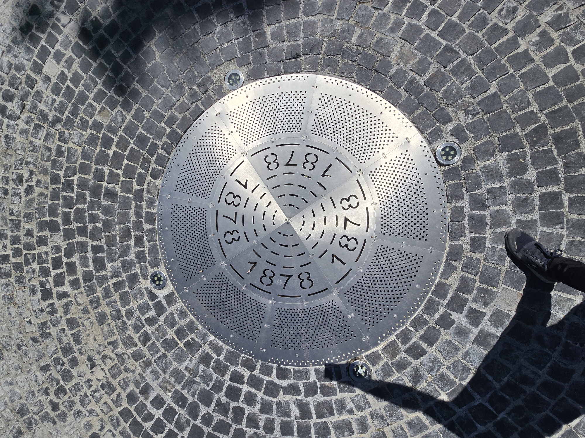 The image shows a circular metal plate set into a cobblestone pavement. The plate is inscribed with the numbers "1878" repeated in a radial pattern. Four small circular lights are embedded in the cobblestone around the edge of the plate. Part of a shoe is visible on the right side of the image. The setting appears to be an urban environment. It is situated in the city of Unknown, Unknown.