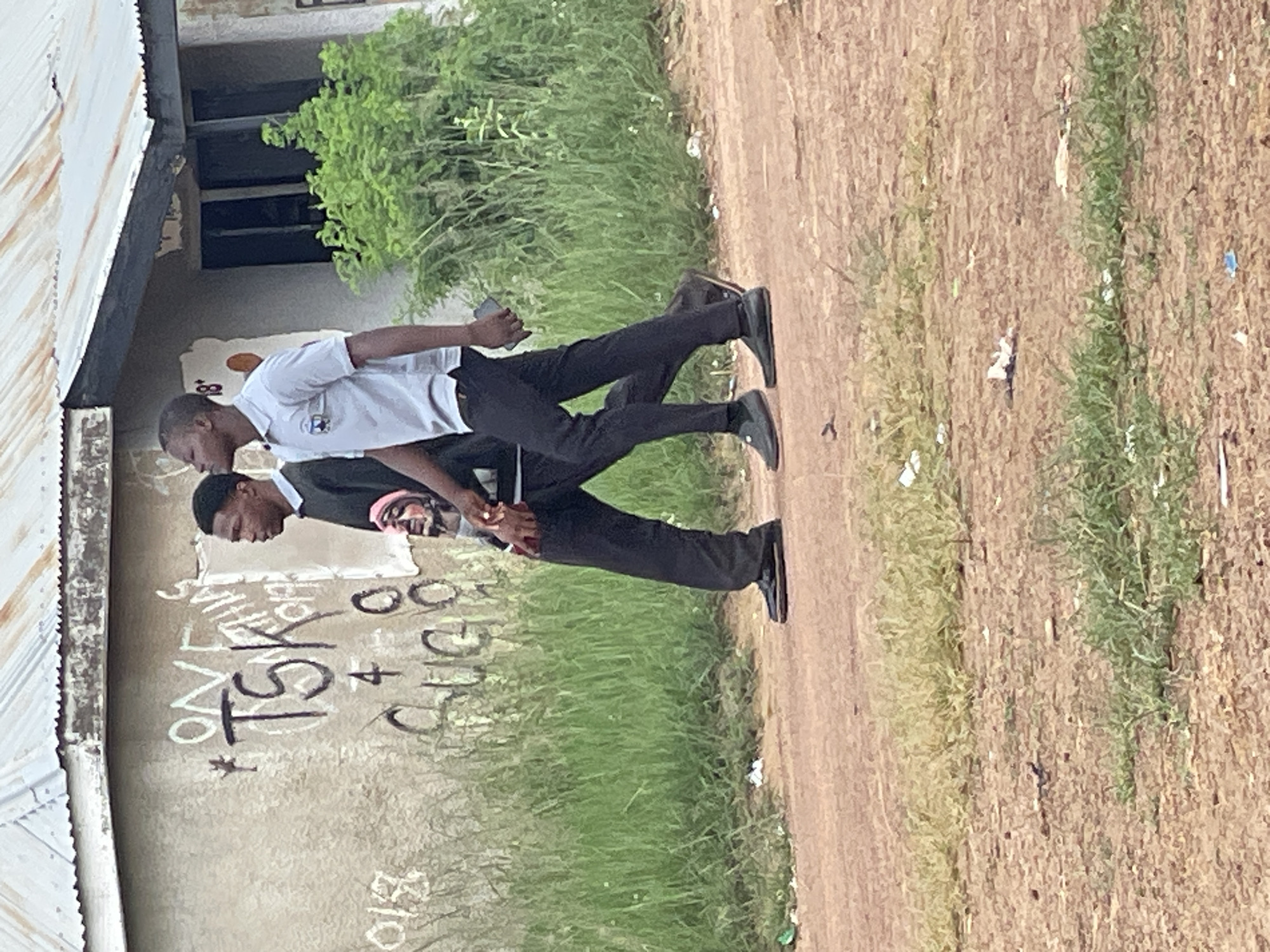In Durumi, Nigeria, two young men are captured in a vertical, close-up shot. One man, wearing a white polo shirt and black trousers, walks ahead, holding a dark object, possibly a phone, in his right hand. Behind him, a second man, dressed in a dark suit with a light pink shirt peeking out, walks in the same direction, his hands clasped behind his back. The wall behind them is adorned with graffiti, including the words "OV", "TSK", and "+". A small white Hello Kitty sticker is also visible on the wall. The scene appears to be outdoors, with a dirt path in the foreground and a patch of green grass and trees in the background, suggesting a daytime setting. The overall mood seems neutral, with no clear indication of strong emotions.